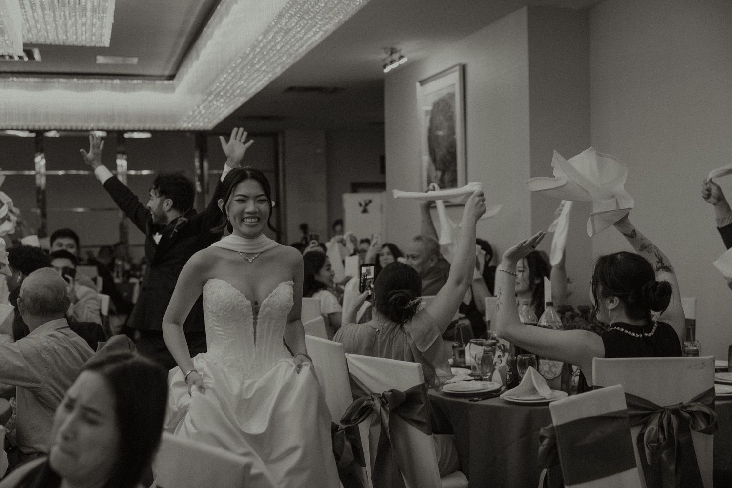 Black and white photo of a smiling bride in a strapless gown entering a wedding reception hall with cheering guests.