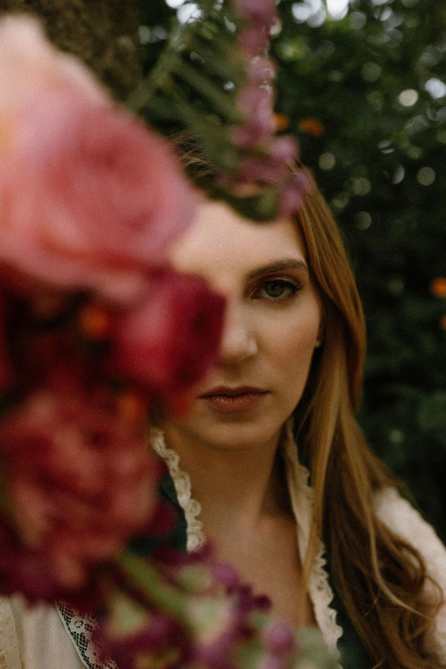 A woman with long brown hair peers over a bouquet of pink and red flowers outdoors.