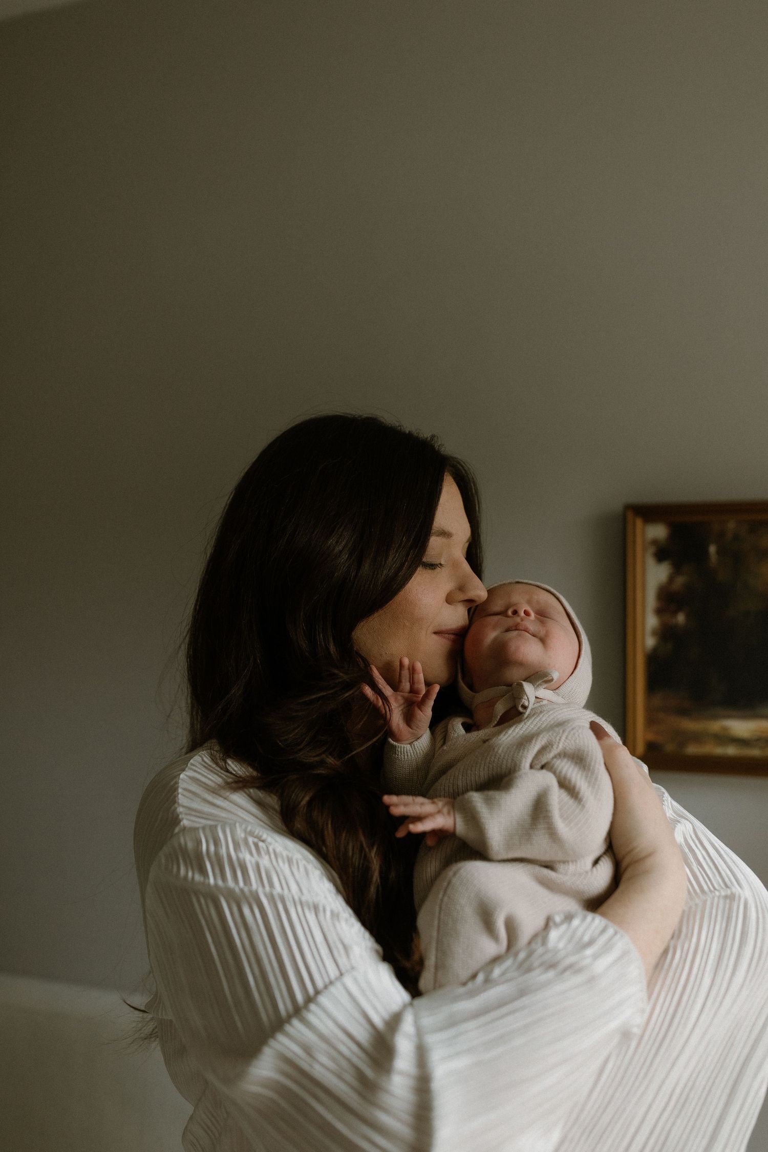 A mother in a white robe tenderly kisses her newborn baby, cradling the infant gently in a softly lit room.