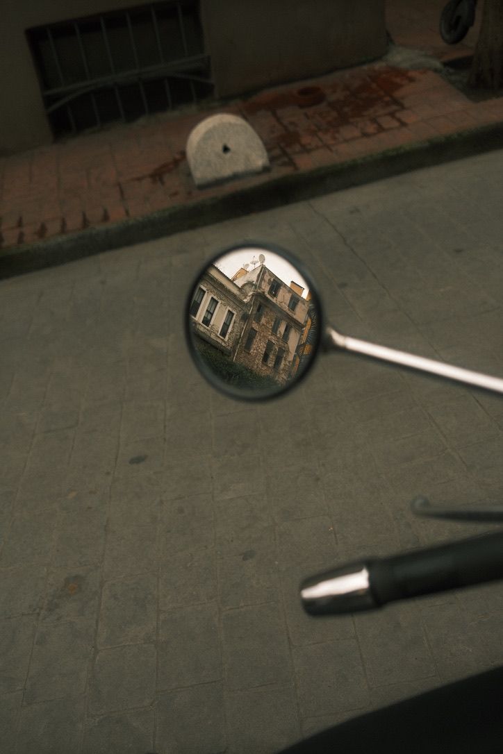 Motorcycle side mirror reflecting an old building facade on a cobblestone street.