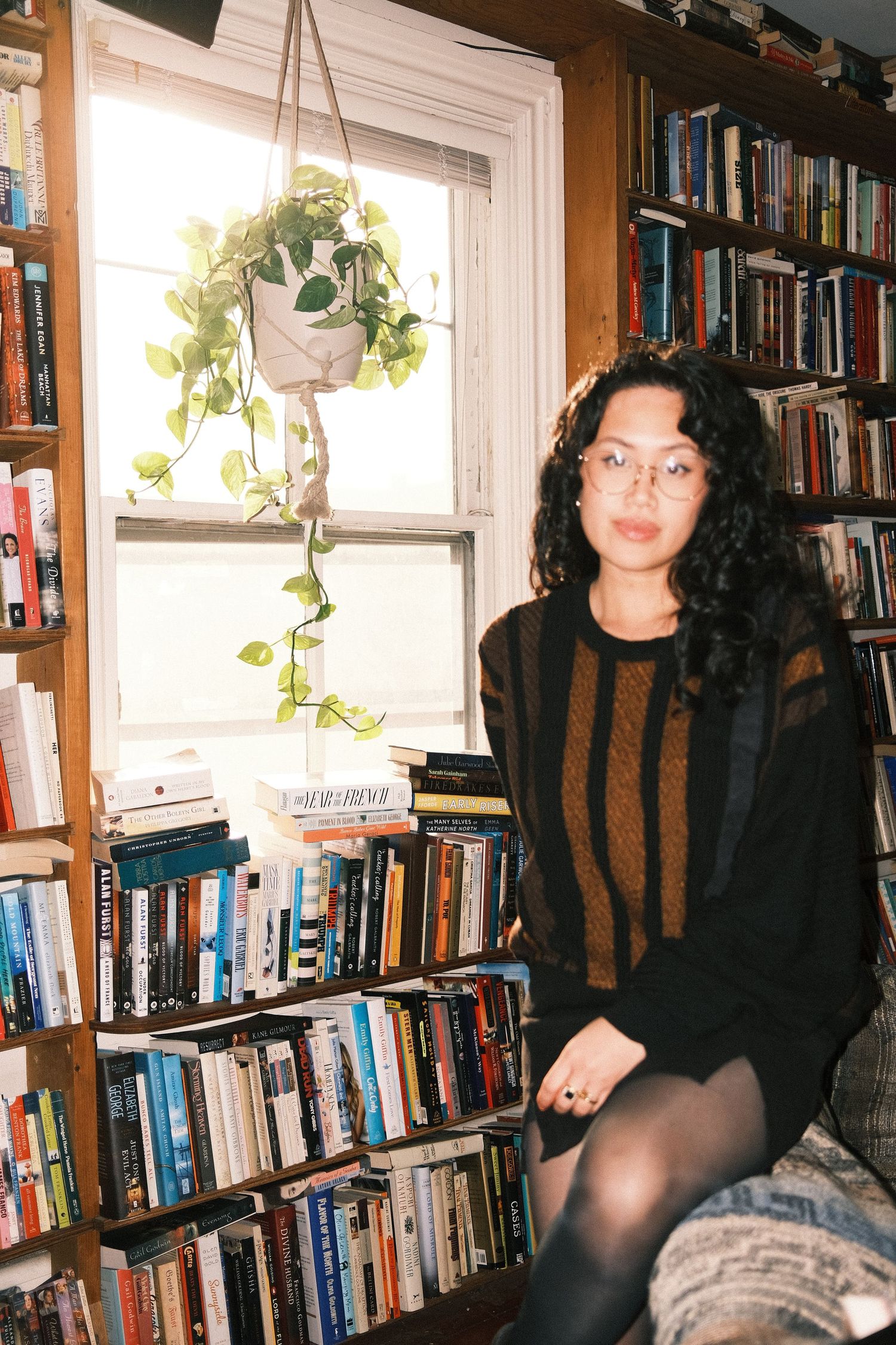 A young woman with curly hair and glasses sits among floor-to-ceiling bookshelves with a hanging pothos plant by a window.