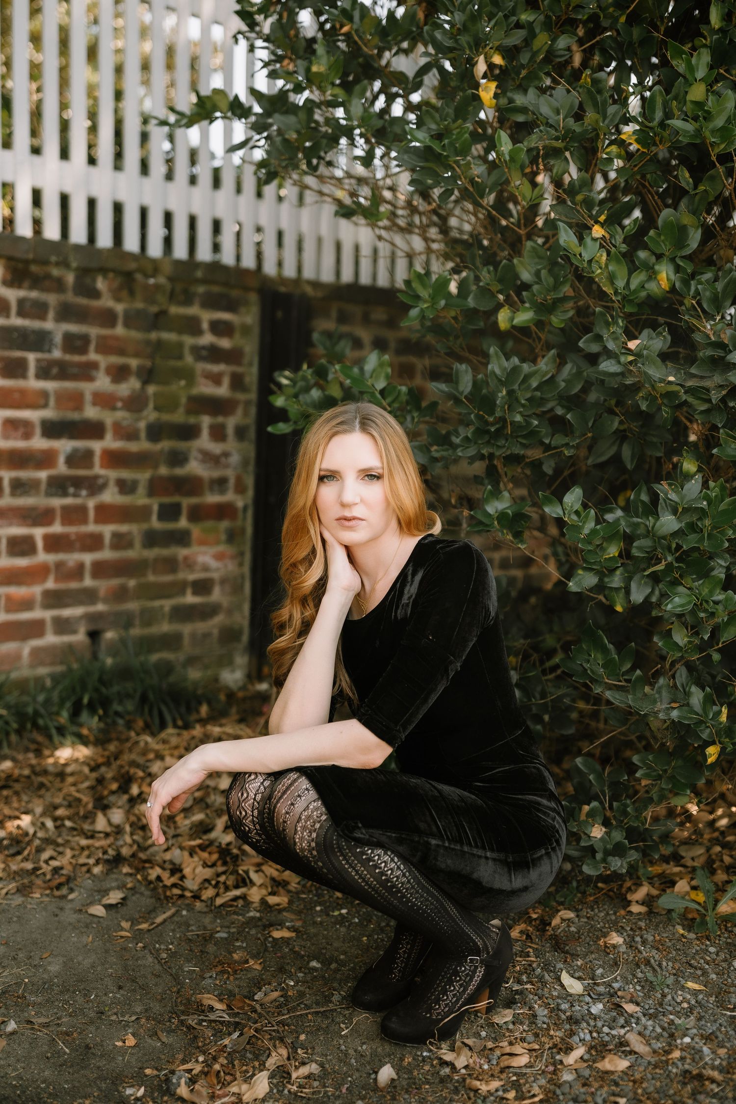 A woman in a black velvet dress and lace tights crouches gracefully beside a brick wall and green shrubs.