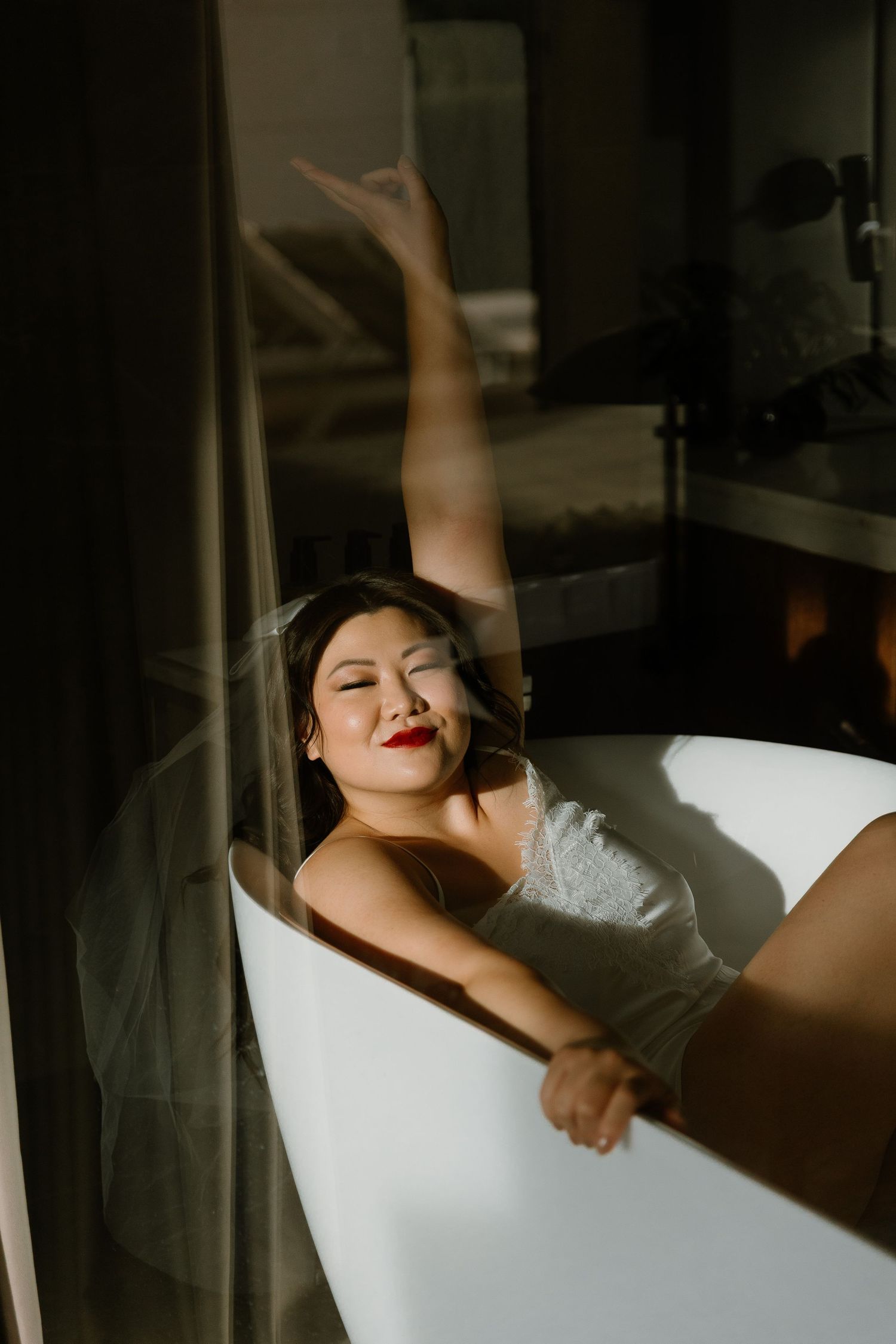 Bride in white lace reclines in freestanding bathtub with arm raised, wearing veil and red lipstick in moody light.