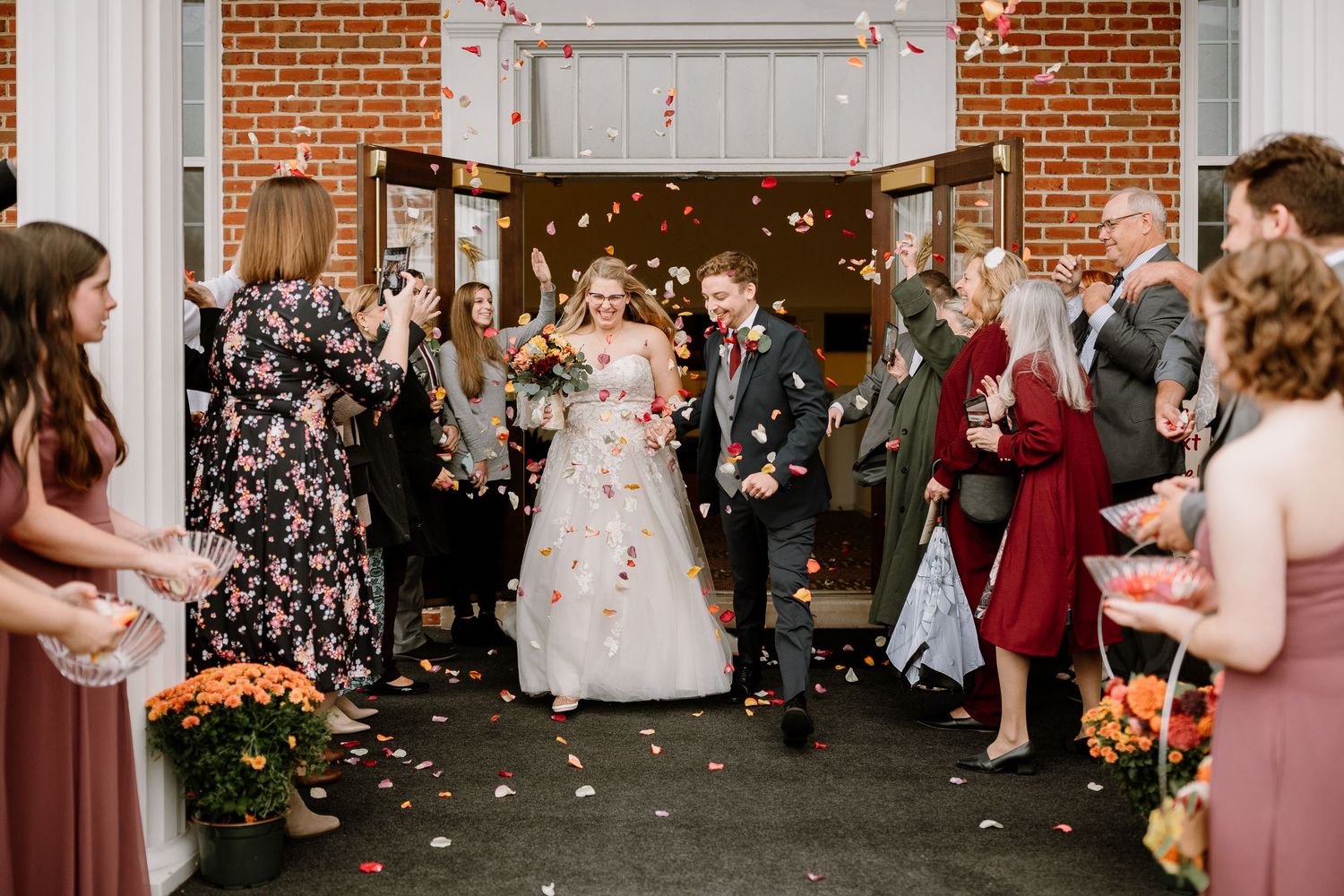 Bride and groom exit wedding venue through flower petal toss with smiling guests lining the path.