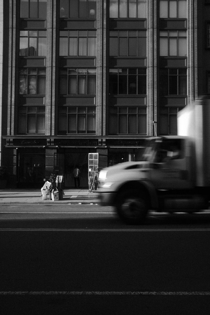 Black and white urban street photo of a blurred moving truck passing a large building with a person sitting on the sidewalk.