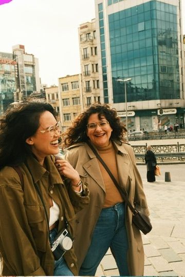 Two women laughing and walking together outdoors in an urban setting, wearing trench coats and glasses.