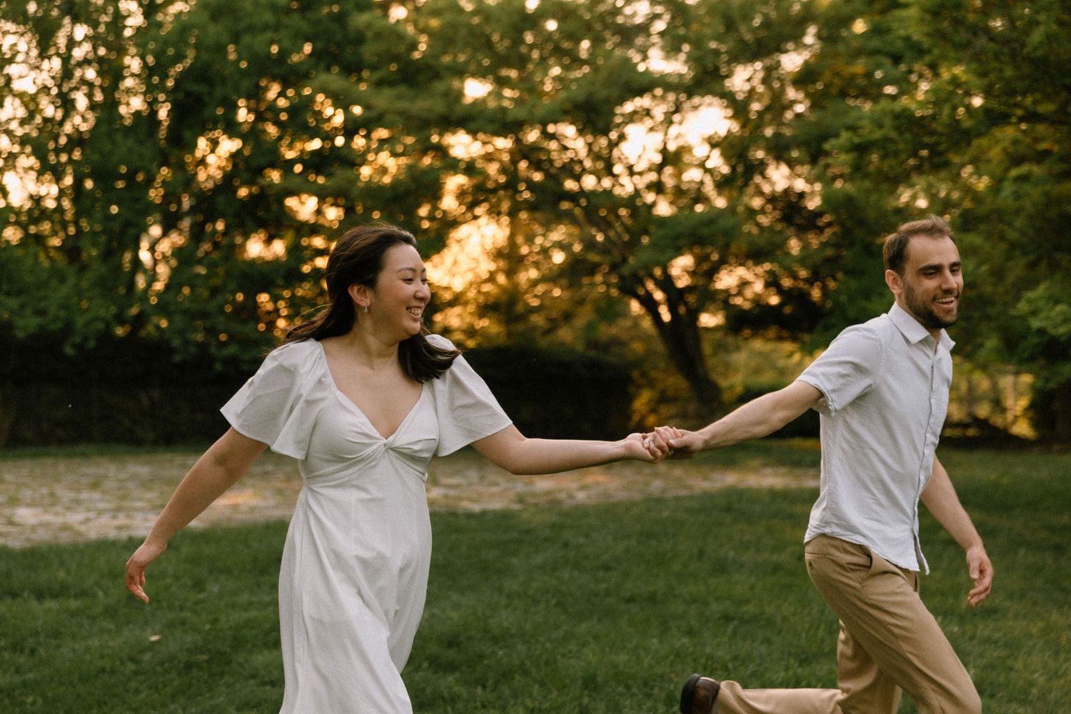 Couple holding hands and laughing while running through a sunlit park at golden hour, woman in white dress.