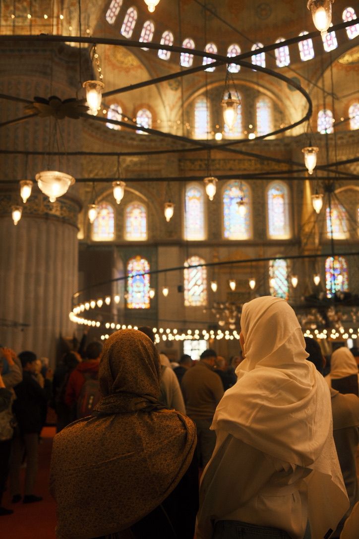 Women in white headscarves praying inside the Blue Mosque with glowing chandeliers and stained glass windows.