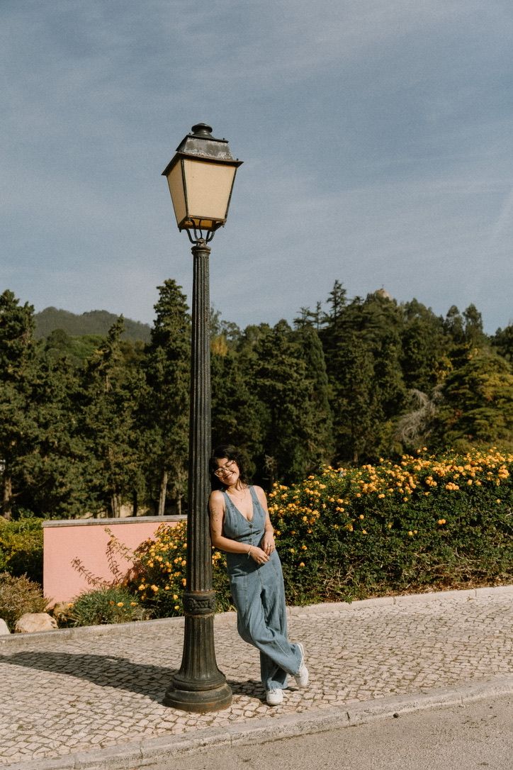 Woman in blue jumpsuit leaning against vintage street lamp, surrounded by lush greenery and yellow flowers on cobblestone path.