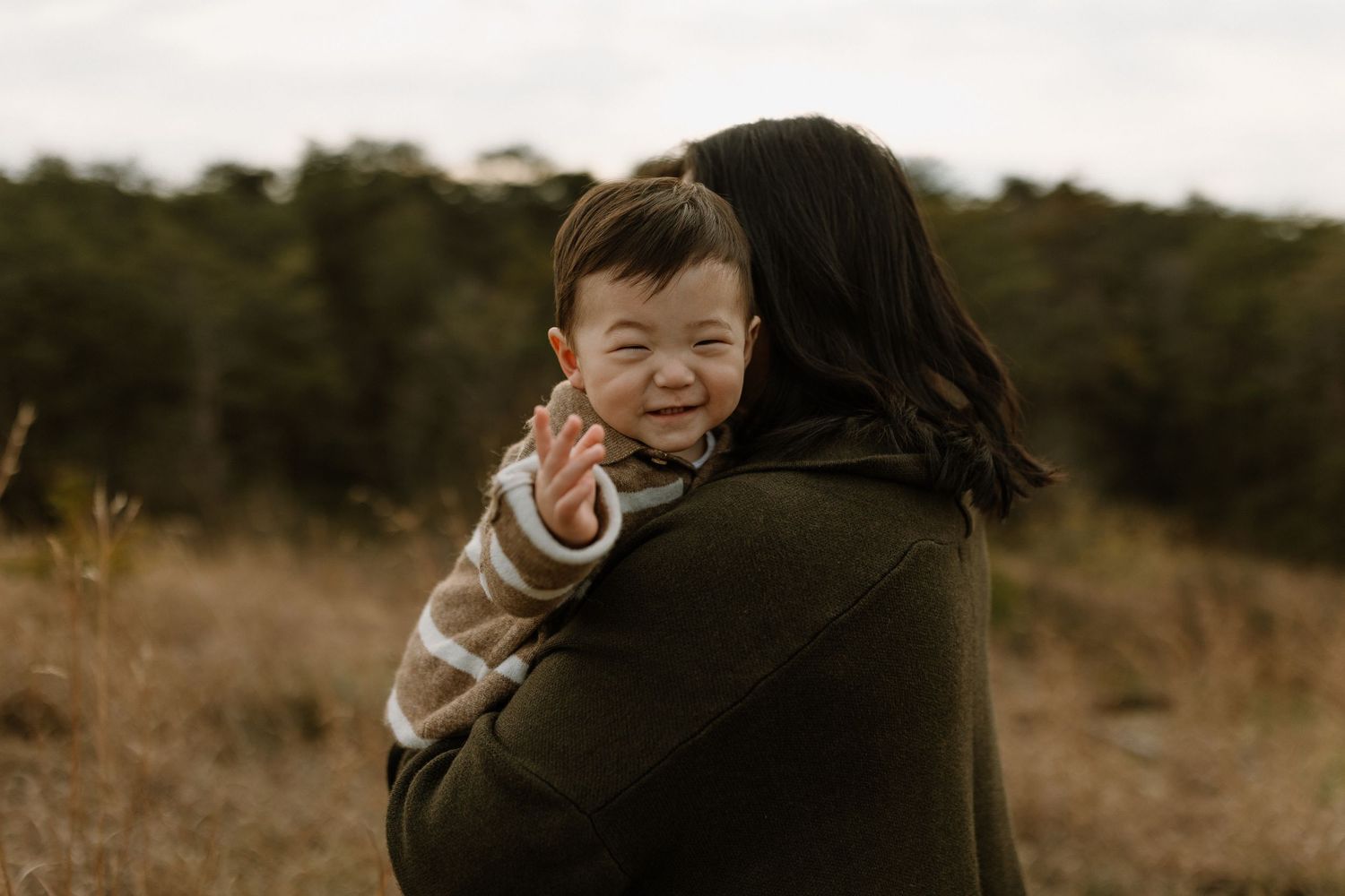 A smiling baby with Down syndrome being held by a woman in a green jacket in a field.