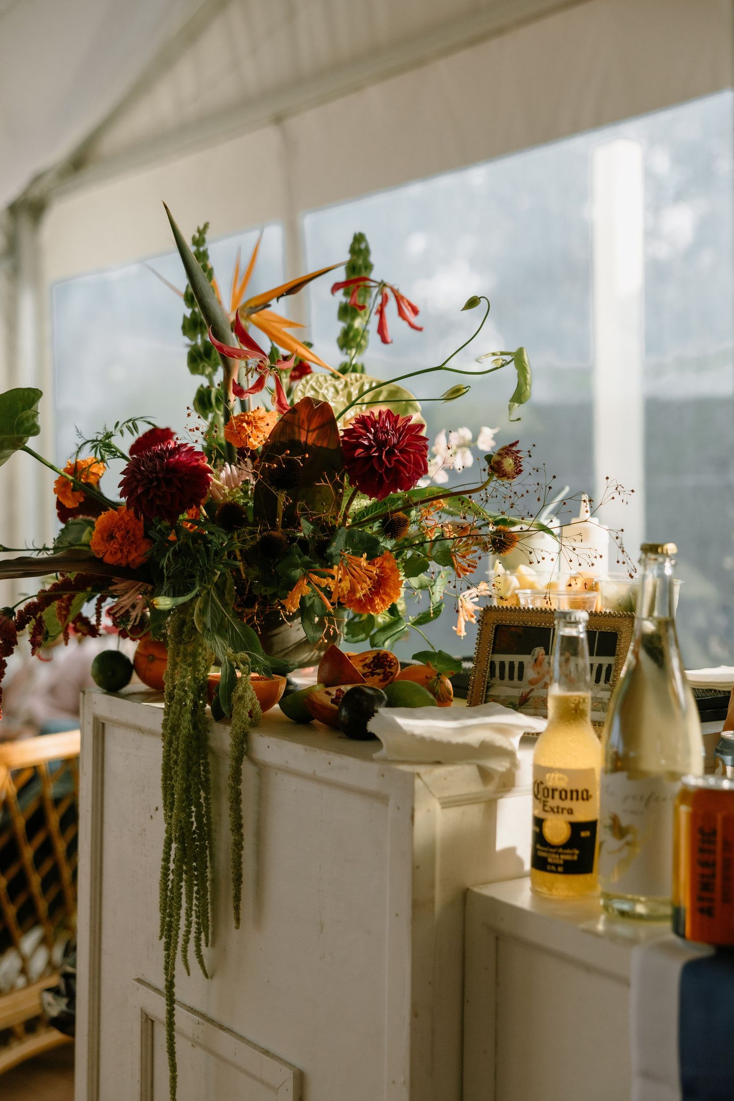 Vibrant floral arrangement with dahlias and birds of paradise beside Corona bottles at an outdoor wedding reception.