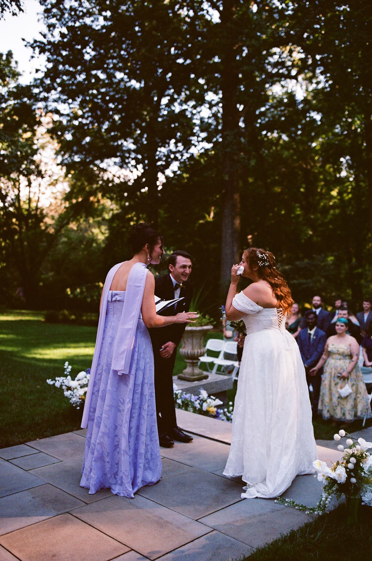 Two brides exchange vows outdoors at a garden wedding ceremony, officiated by a man, surrounded by lush greenery.