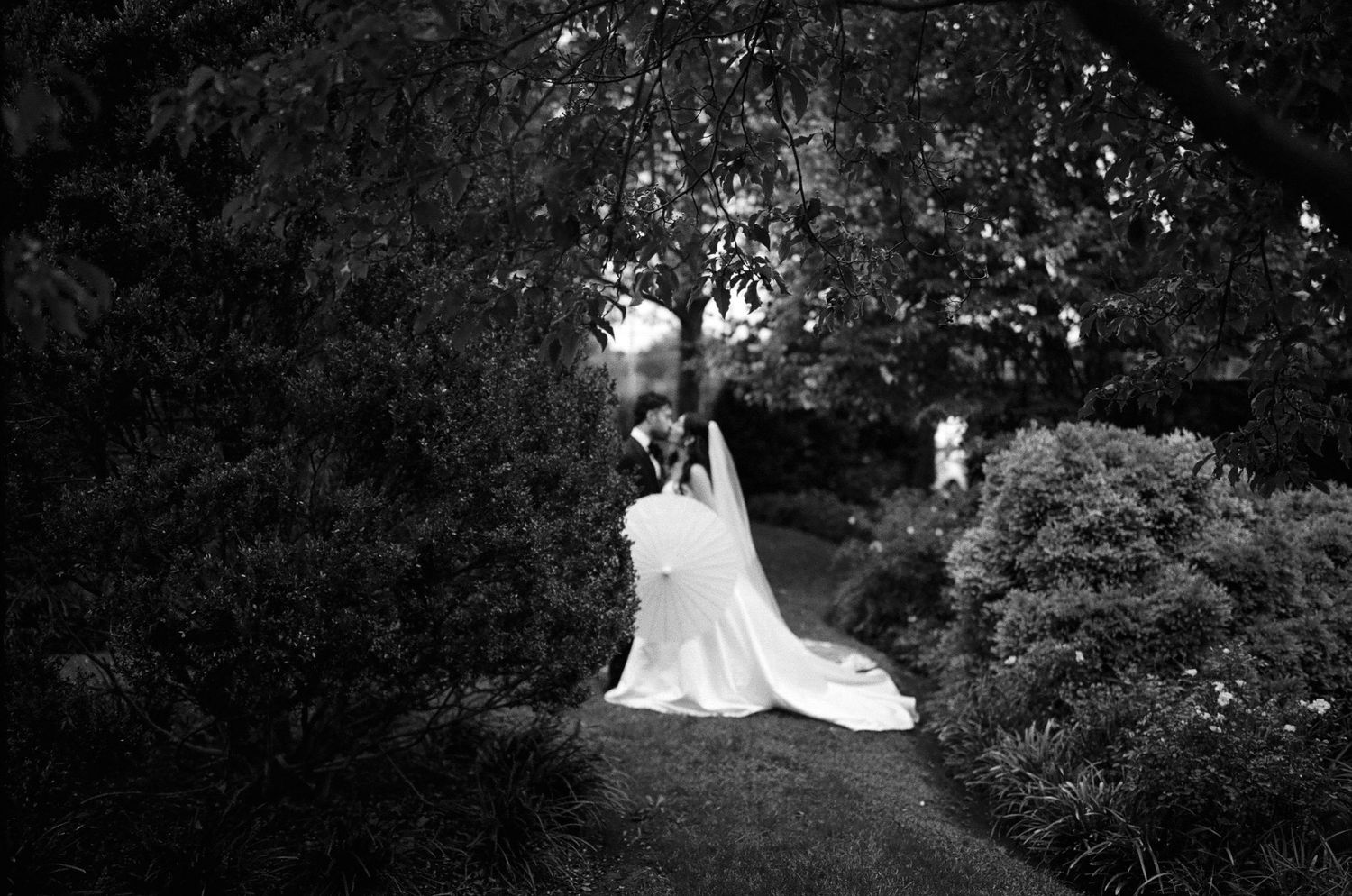 Black and white photo of a bride in a flowing white gown and veil standing in a lush garden pathway.