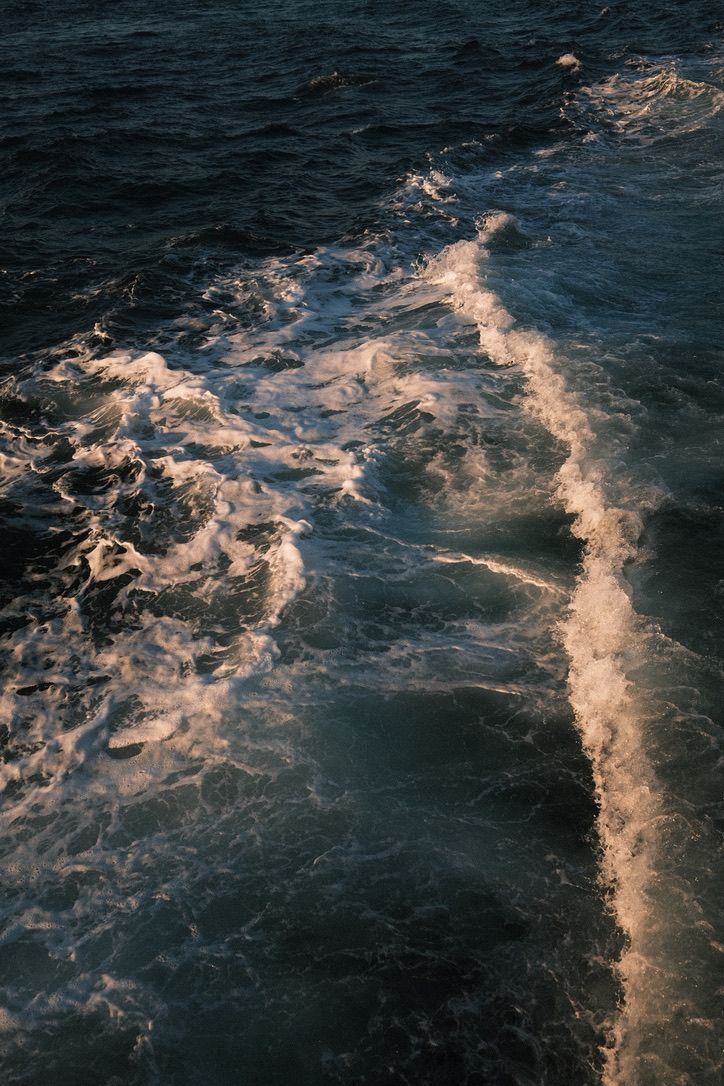 Dark ocean waves with white foam wake trail glowing in warm sunset light, aerial view of turbulent sea water.