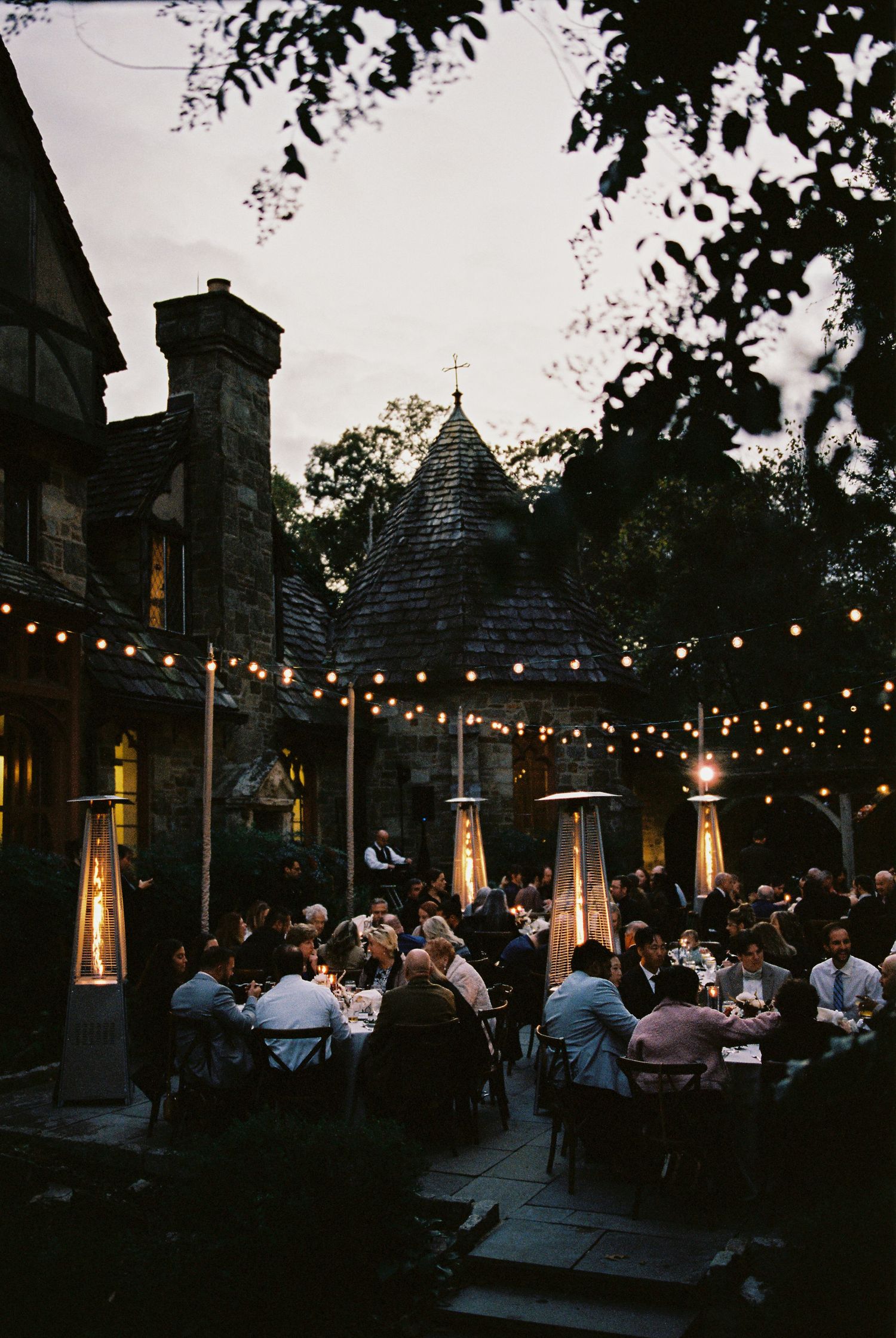 Outdoor evening wedding reception with string lights, patio heaters, and guests dining near a charming stone building at dusk.