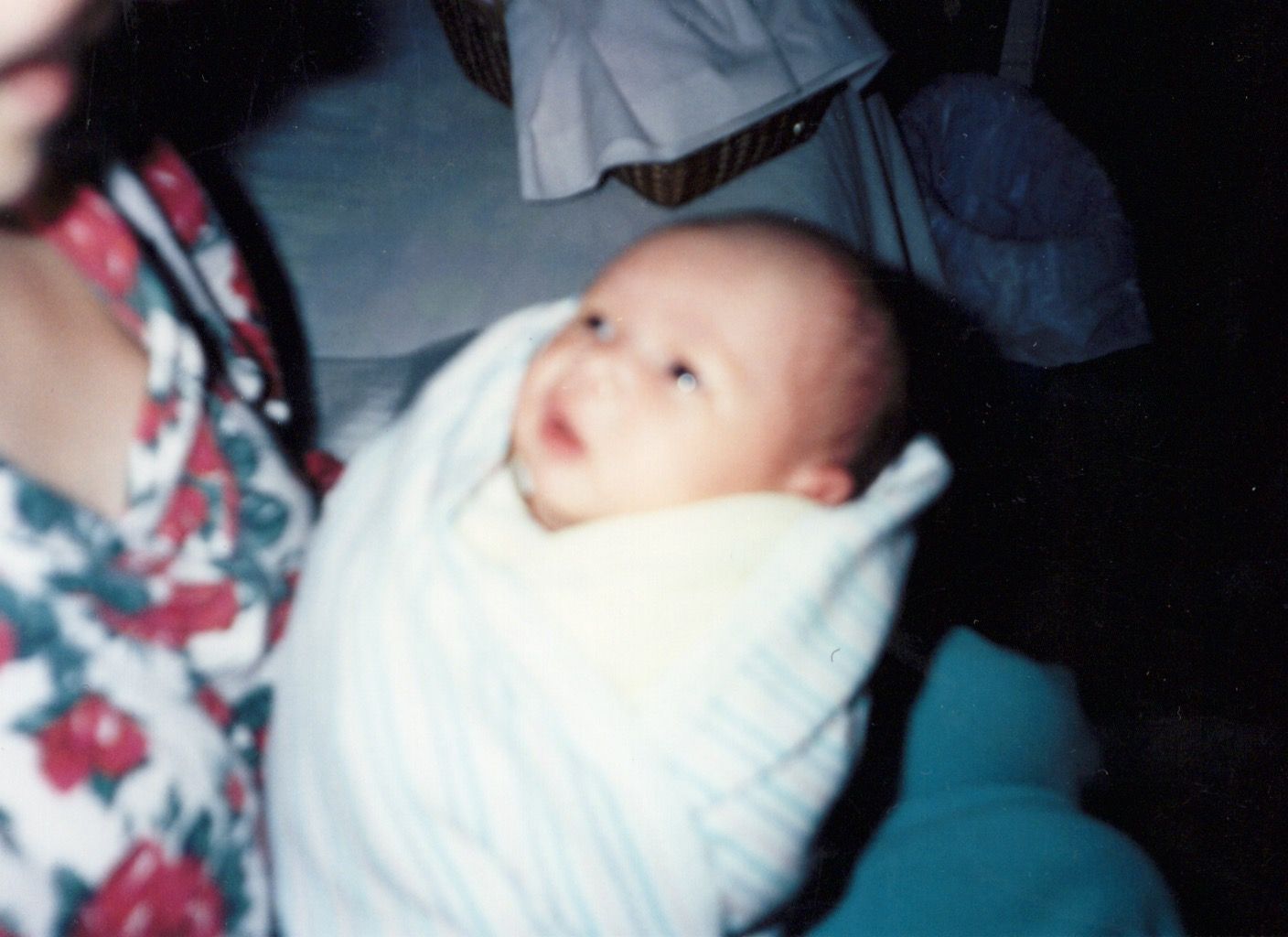 A newborn baby swaddled in a white blanket being held by a person in a floral dress in a dimly lit room.