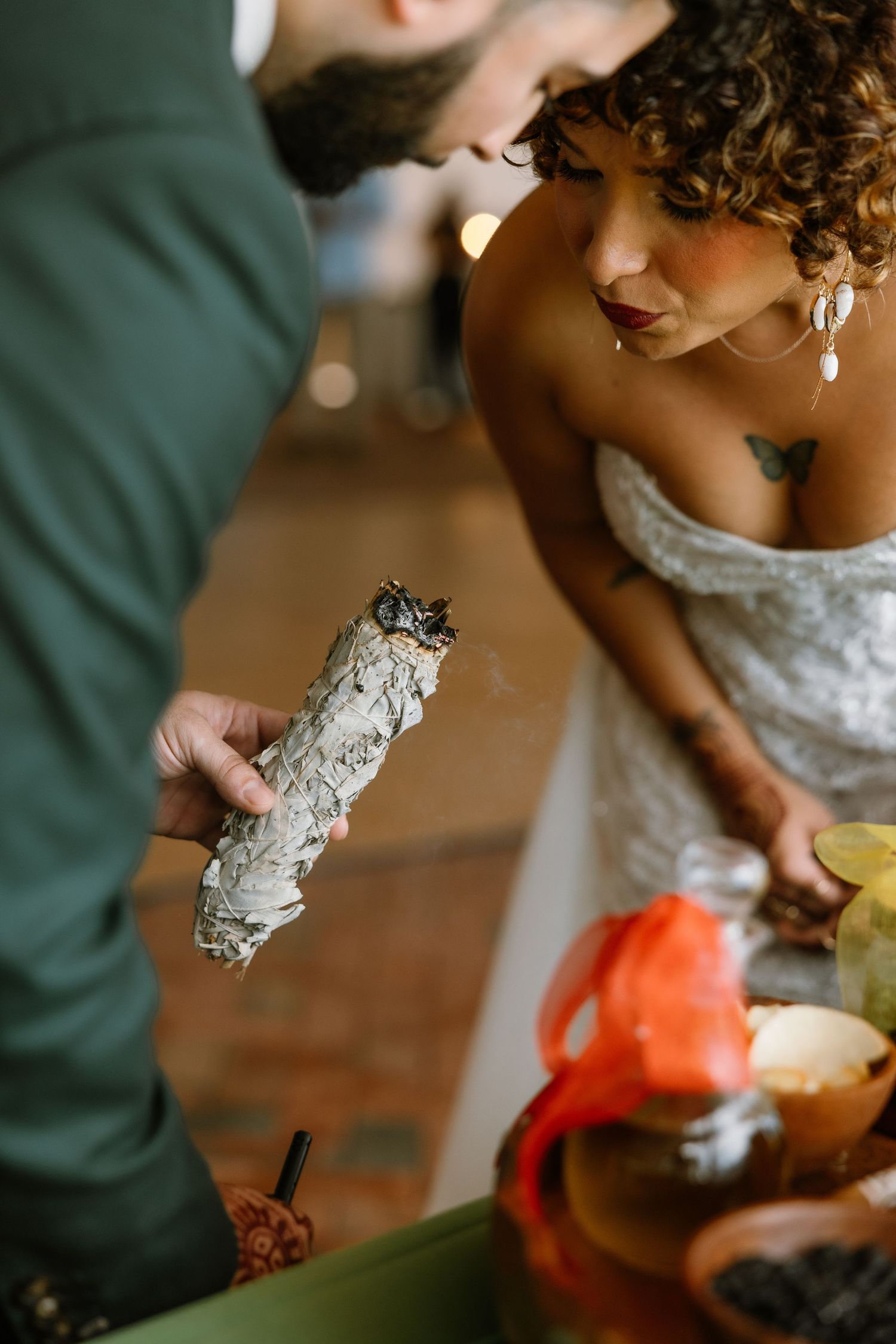 Bride in lace dress watches groom hold a white sage smudge stick during a boho wedding ceremony ritual.