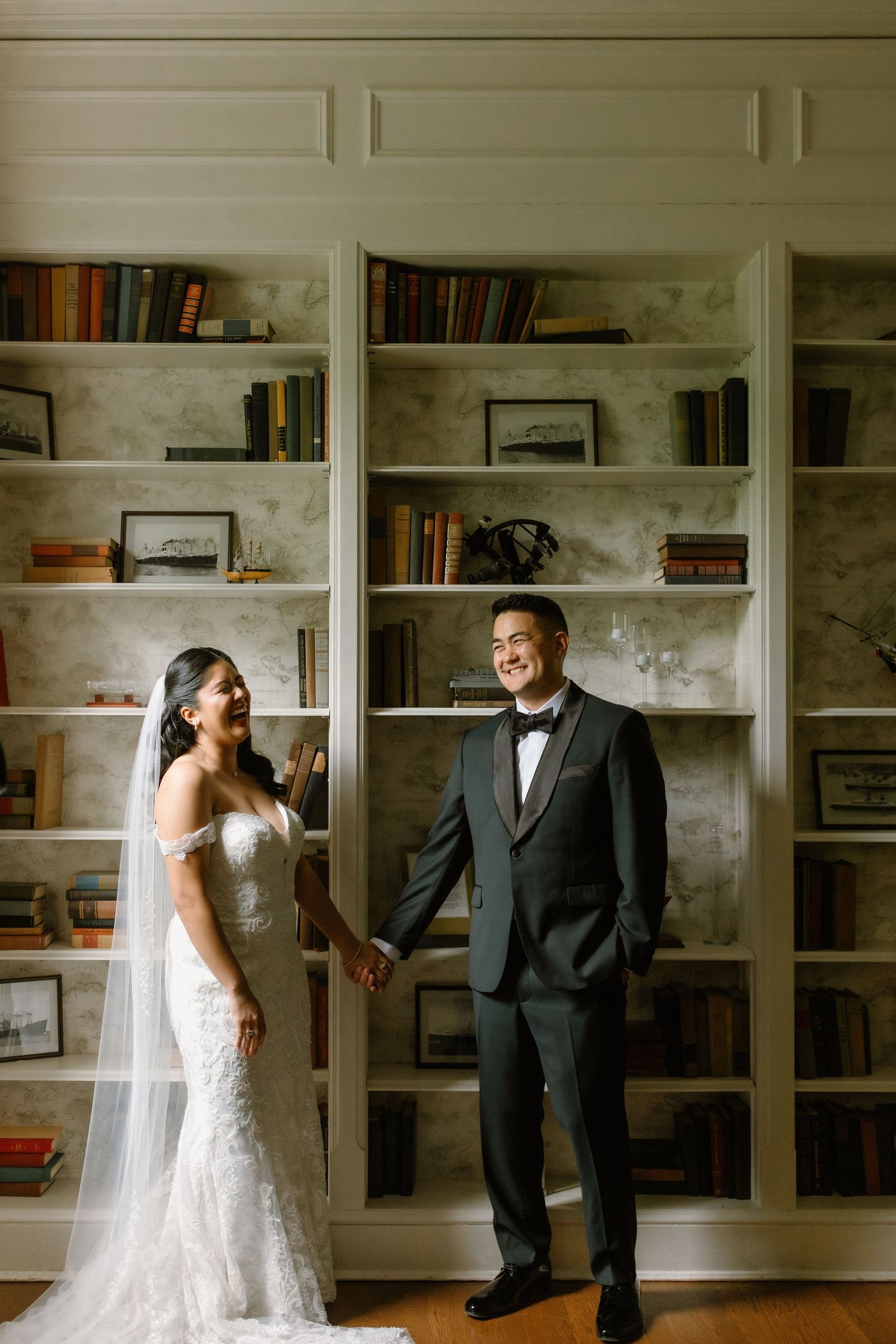 Bride and groom hold hands smiling in front of elegant bookshelf backdrop, bride in lace gown with veil, groom in black tuxedo.