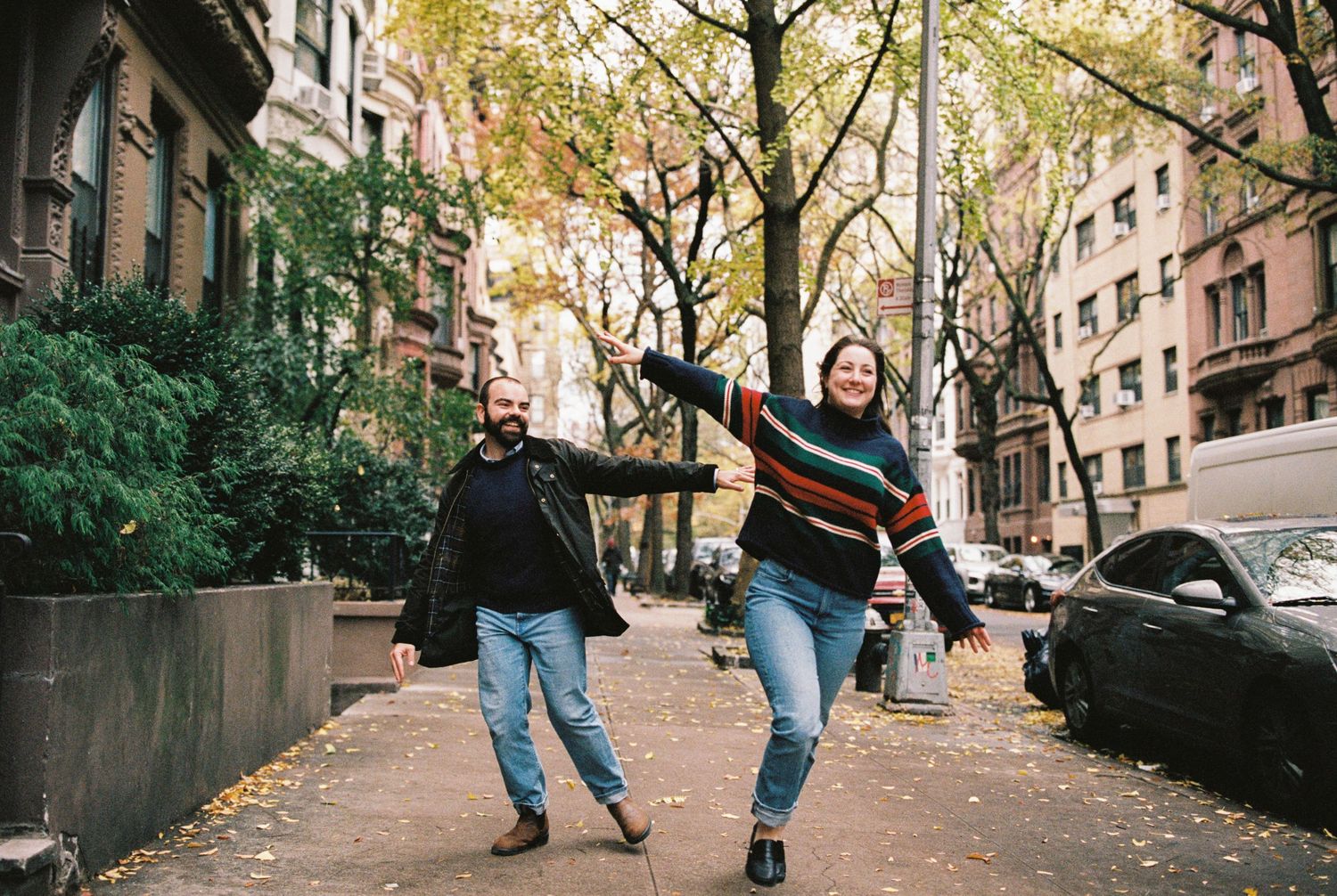 Happy couple runs playfully down a tree-lined urban sidewalk during fall engagement photo session.