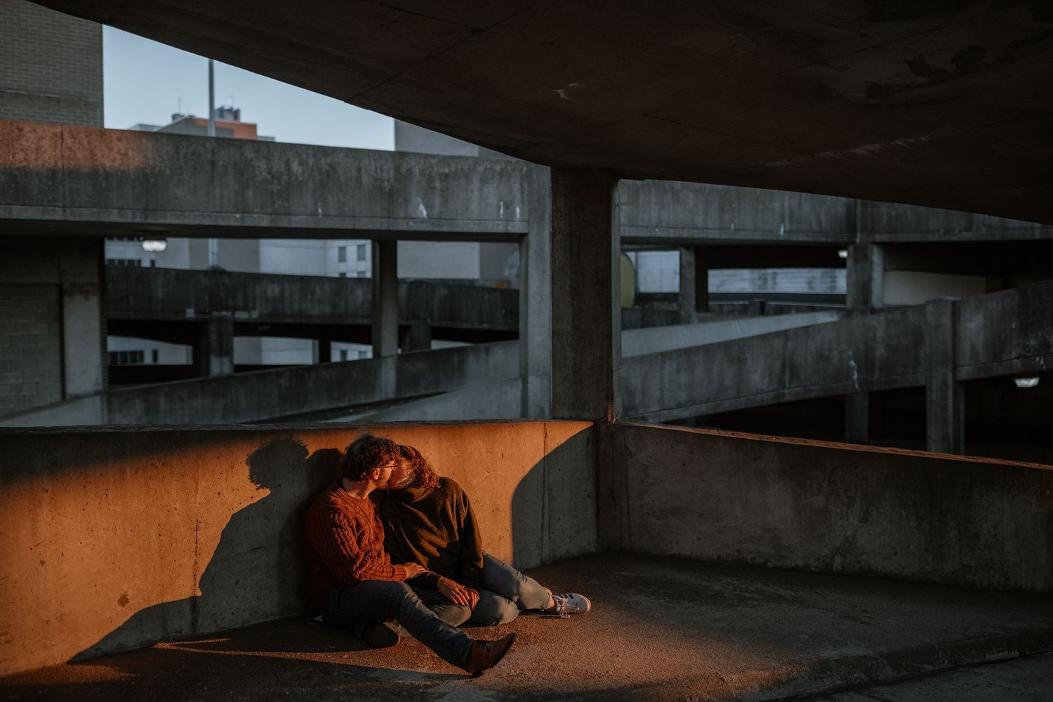 Couple sitting together under a concrete parking structure at dusk, bathed in warm golden light.