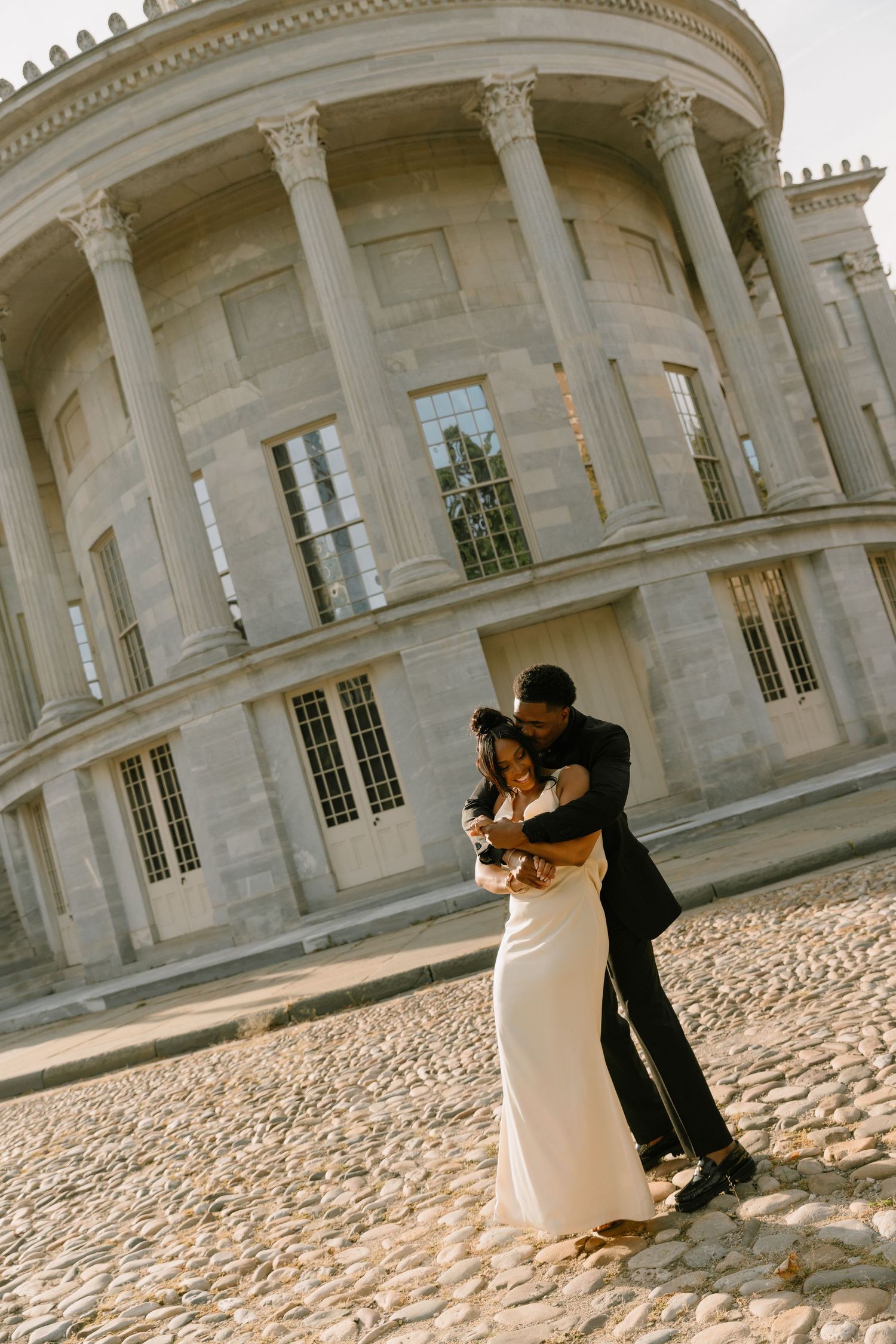 Couple dancing in wedding attire on cobblestone plaza before grand neoclassical building with tall columns.