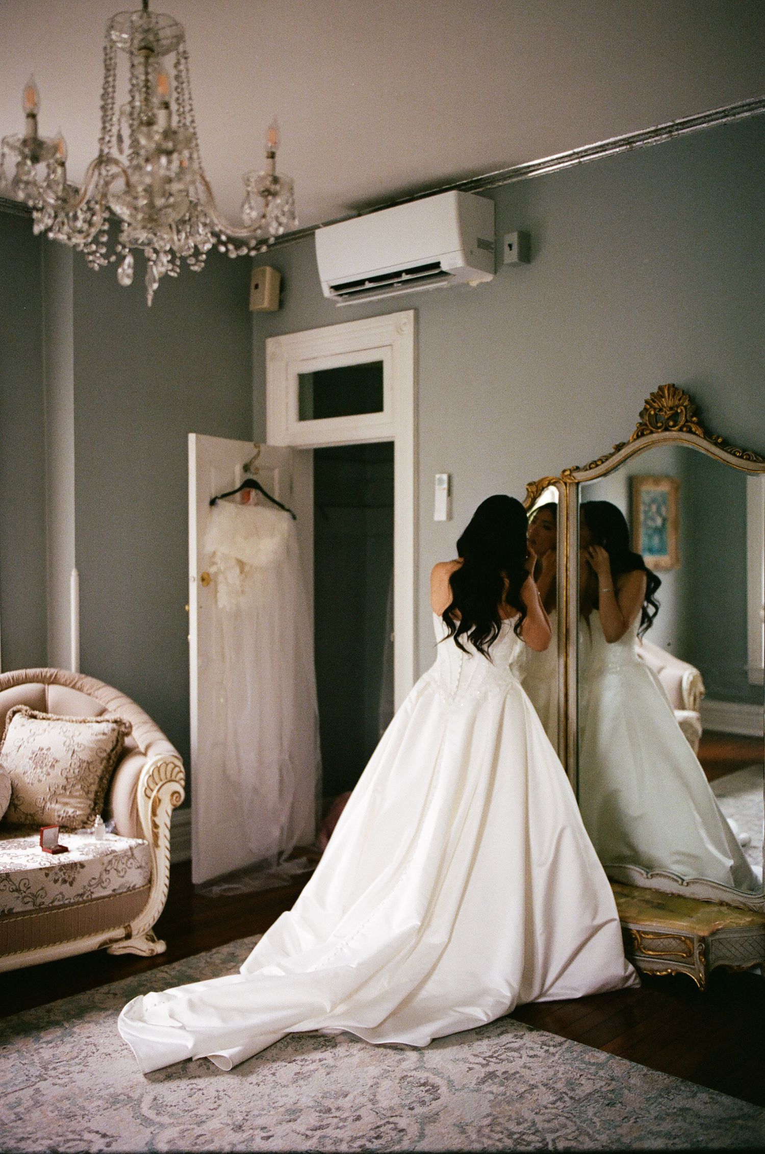 Bride in a white ballgown wedding dress stands before a gilded mirror in an elegant bridal suite with crystal chandelier.