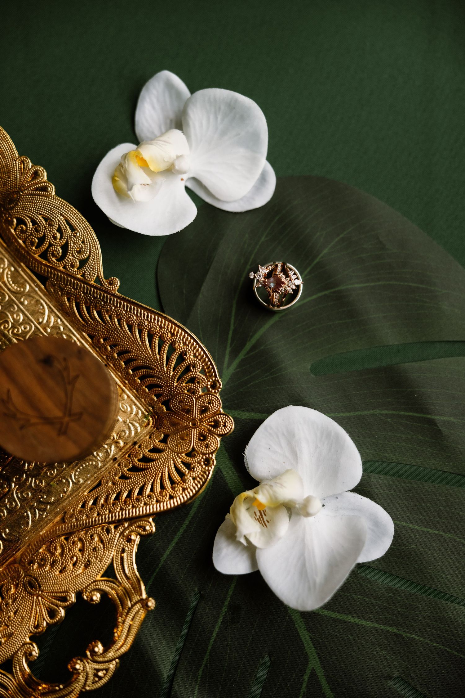 Gold ornate tray with white orchids and monstera leaf on dark green background, elegant tropical flat lay.