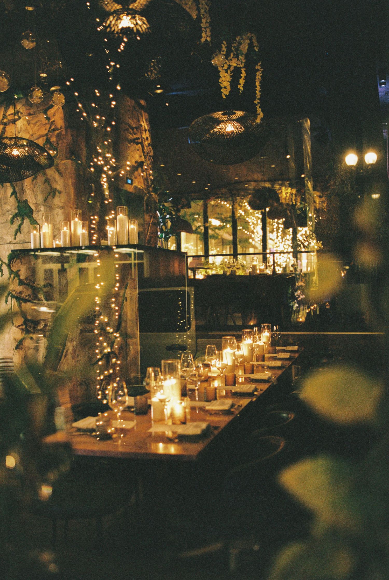 Romantic candlelit restaurant with fairy lights, long wooden dining table, and lush greenery in a cozy cave-like setting.