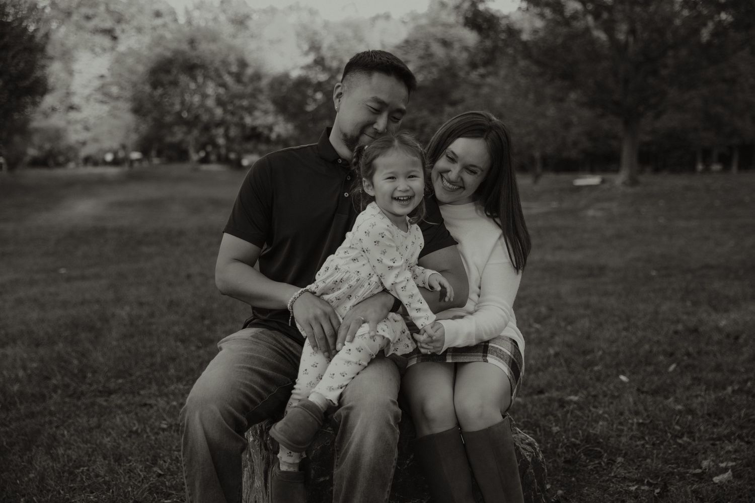 Black and white family portrait of parents smiling with toddler daughter outdoors in a park setting.