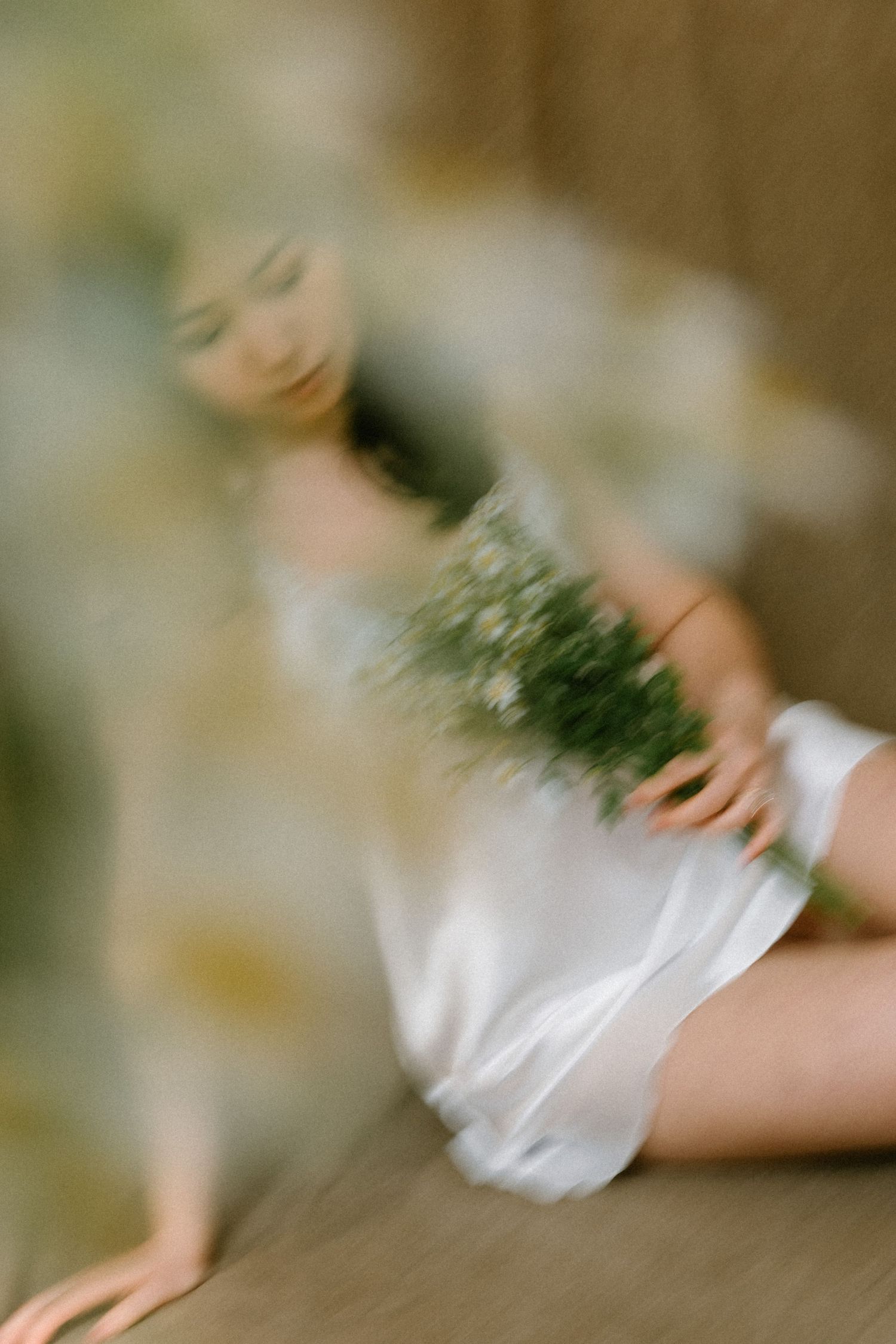 A woman in a white slip dress sits gracefully holding a bouquet of wildflowers, captured in a dreamy, soft-focus artistic photo.