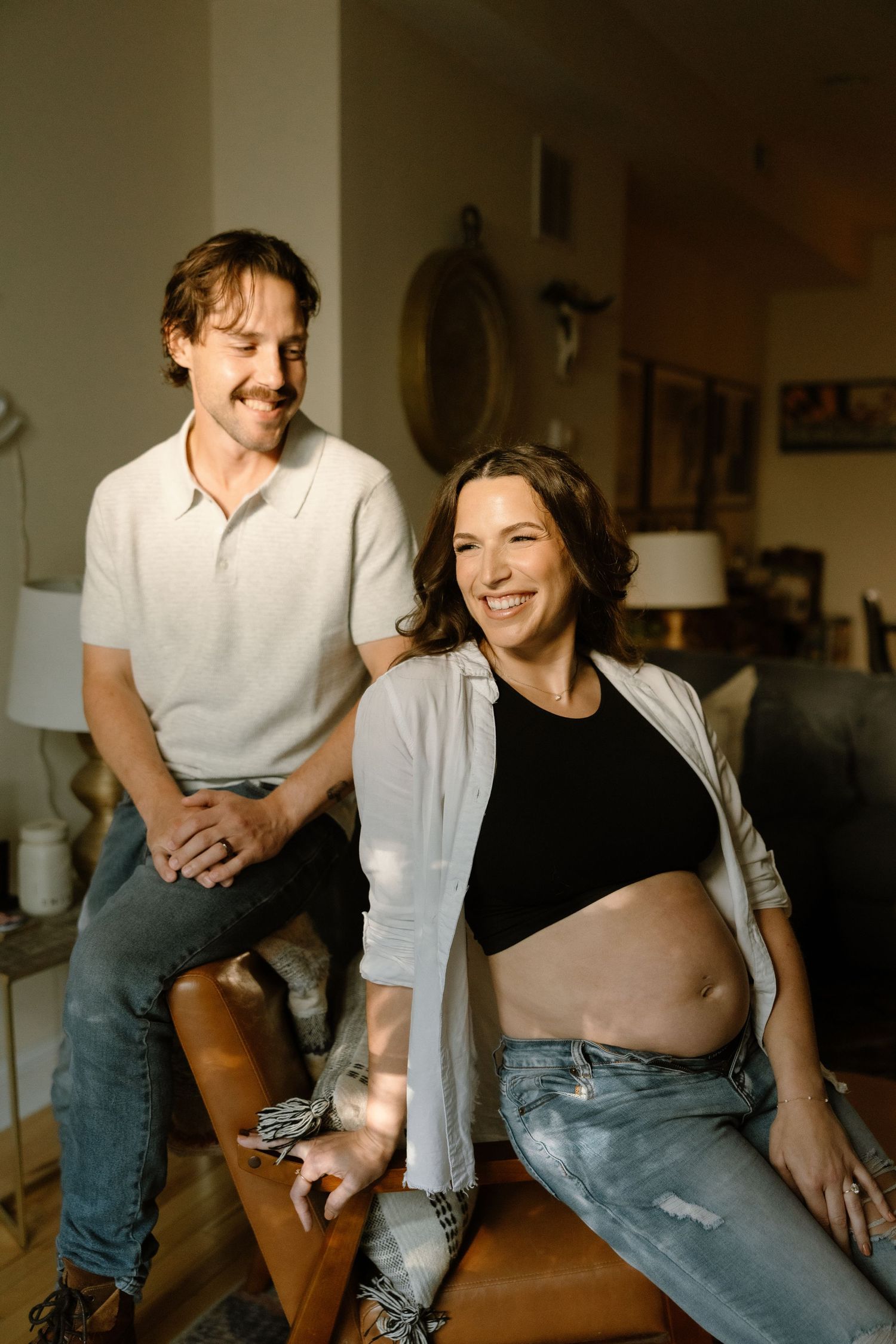 A smiling pregnant woman in jeans and crop top sits with her partner in a warmly lit living room.