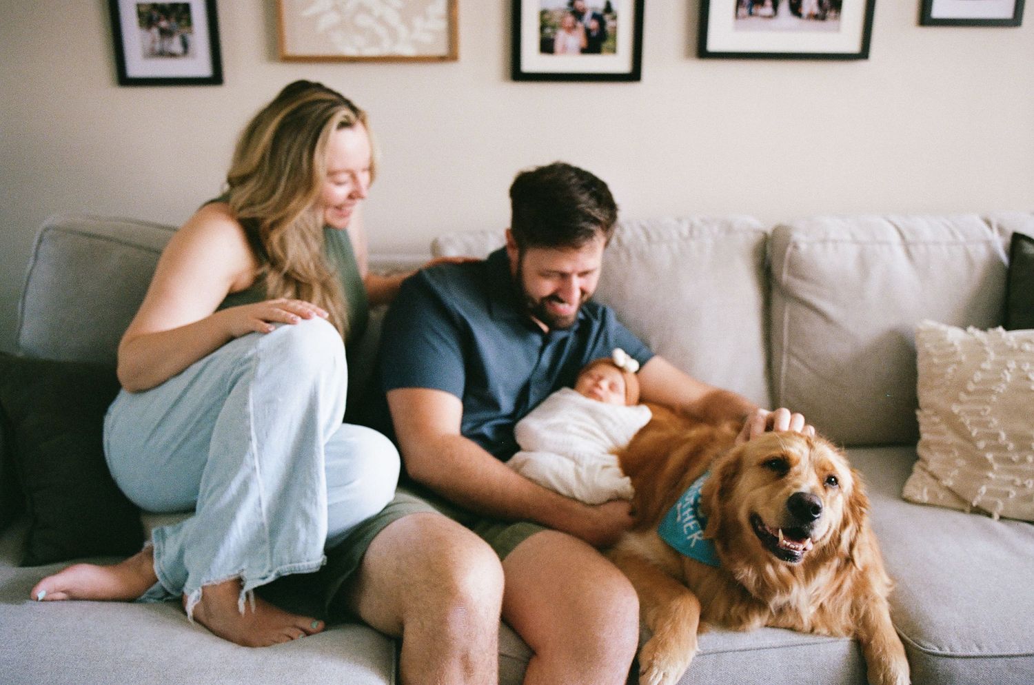 Happy family with newborn baby and golden retriever dog sitting together on a cozy gray couch at home.