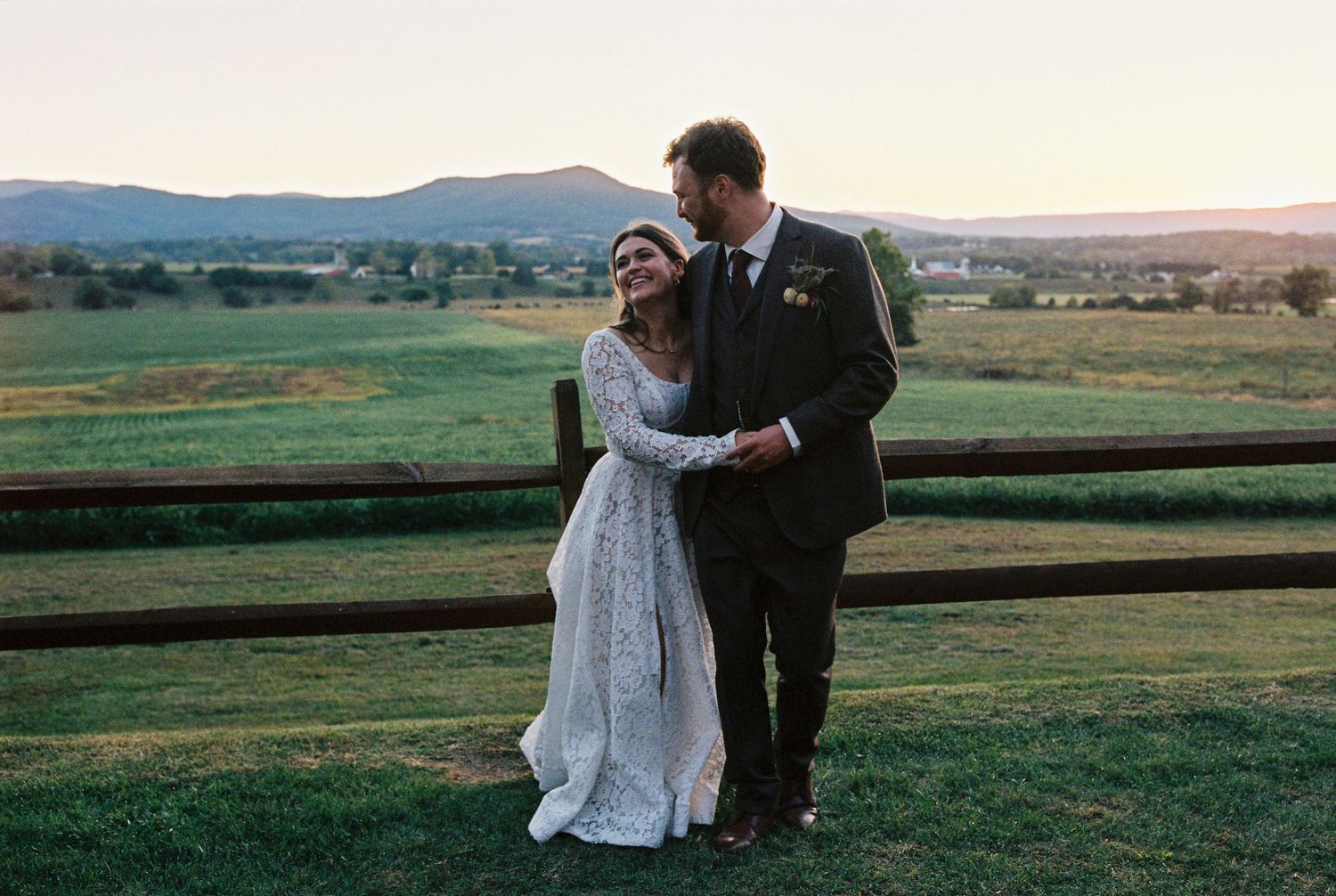 Bride and groom embracing at sunset with scenic mountain views and green fields at an outdoor wedding venue.