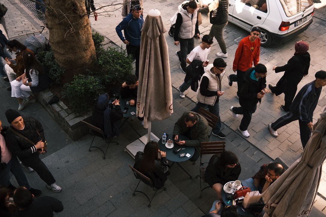 Aerial view of people sitting at outdoor cafe tables on a cobblestone plaza with pedestrians walking by.