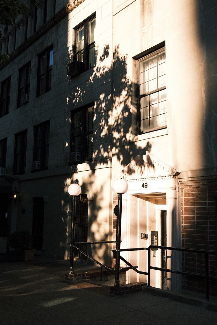Sunlit building entrance at number 49 with tree shadows cast on facade and glowing street lamps on urban sidewalk.