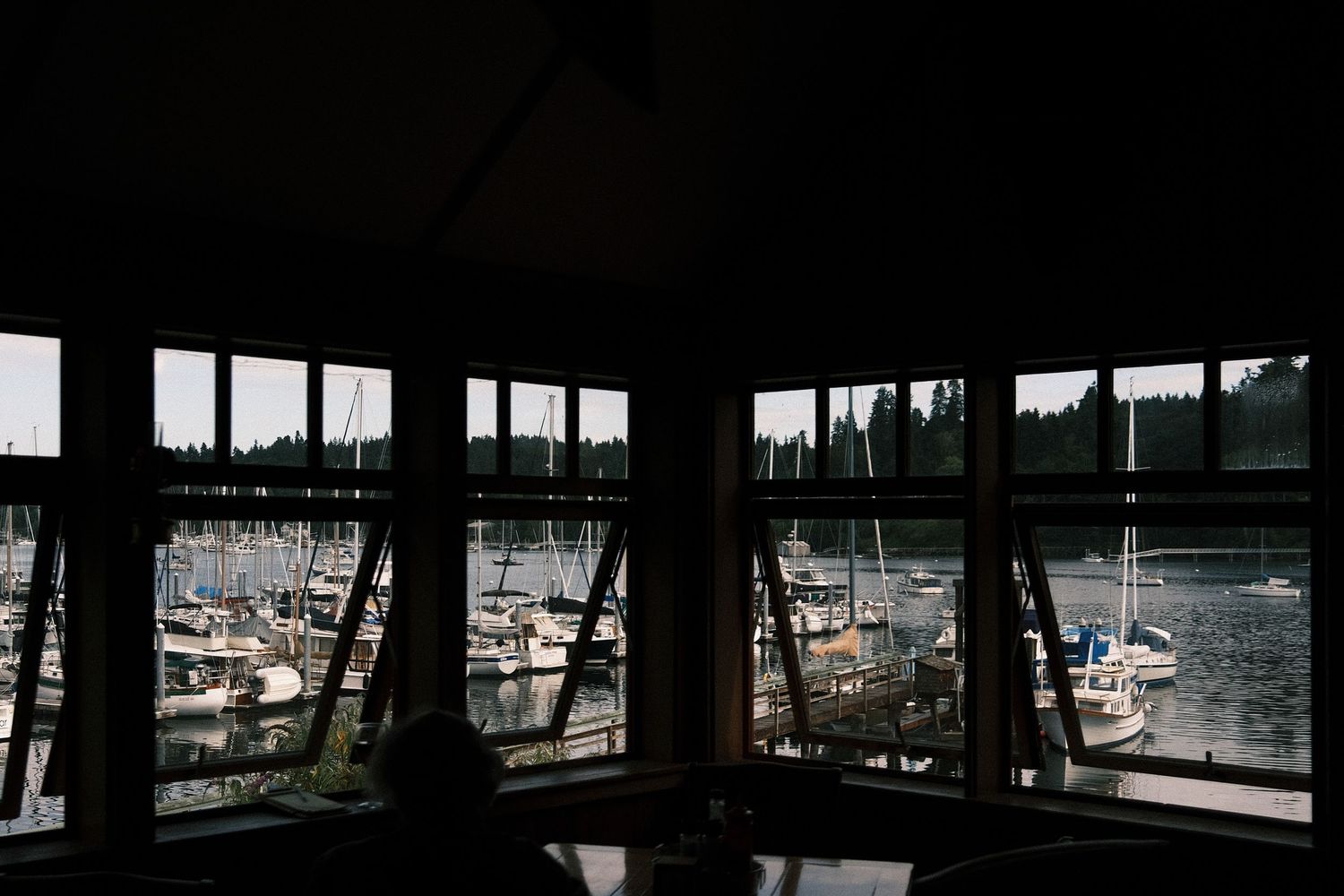 View from inside a marina building with large windows overlooking sailboats docked in a calm harbor surrounded by trees.