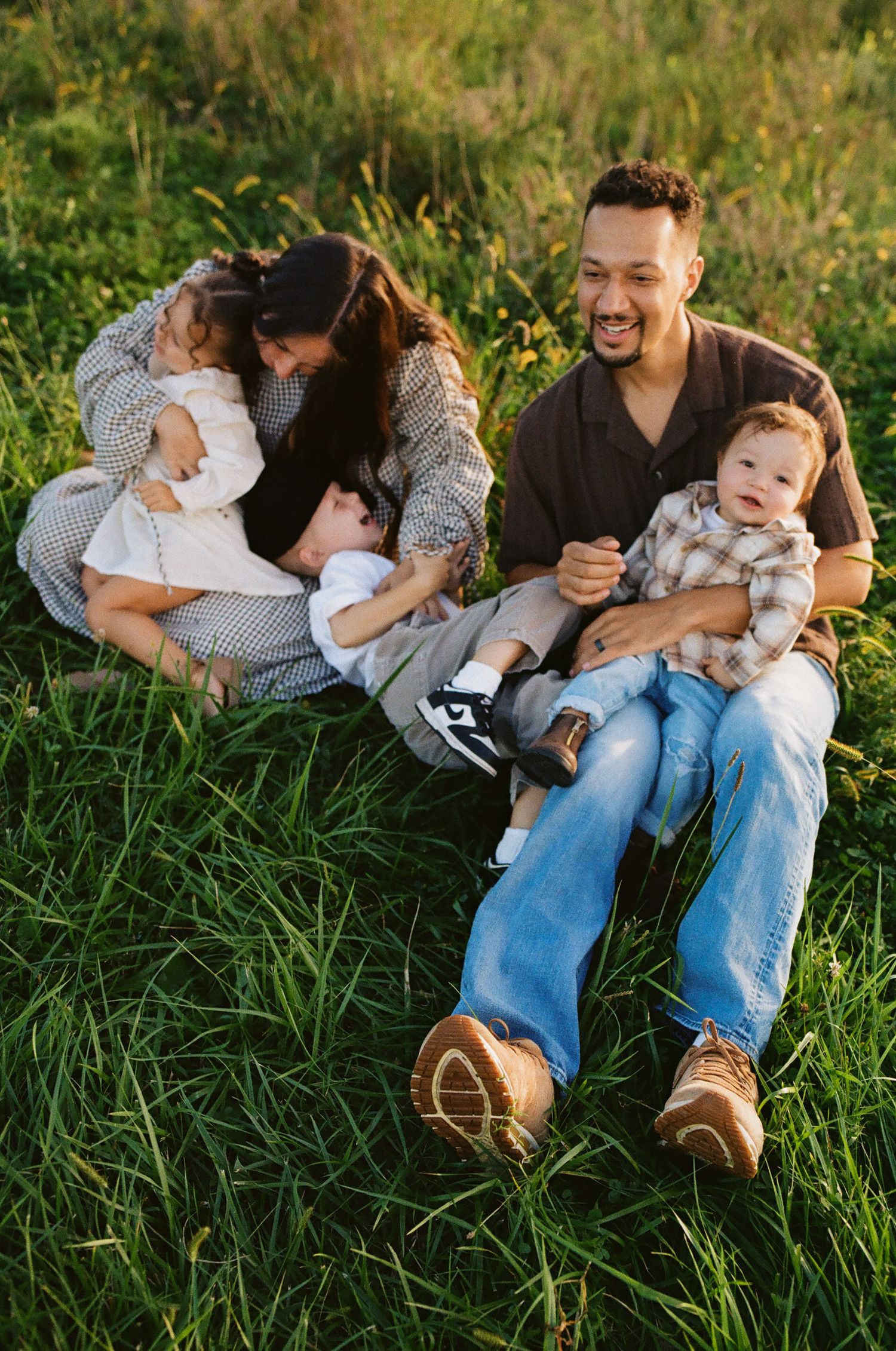 Family of four sitting in lush green grass, parents smiling and cuddling two young children outdoors.