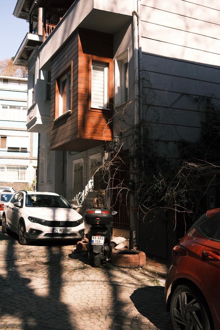 Modern residential building with wood-paneled balcony, parked cars and motorcycle on a cobblestone street in sunlight.