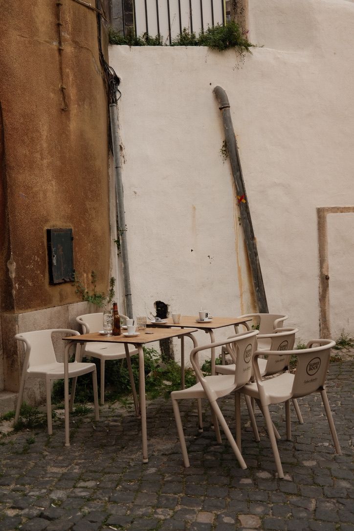 Rustic outdoor cafe with wooden tables and white chairs on cobblestone street beside weathered walls.