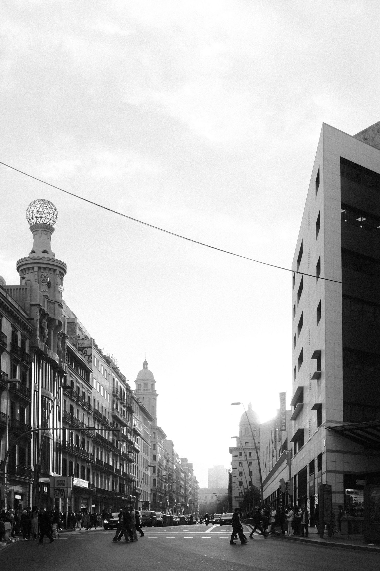 Black and white urban street scene with pedestrians, historic buildings, and a decorative tower under a cloudy sky.