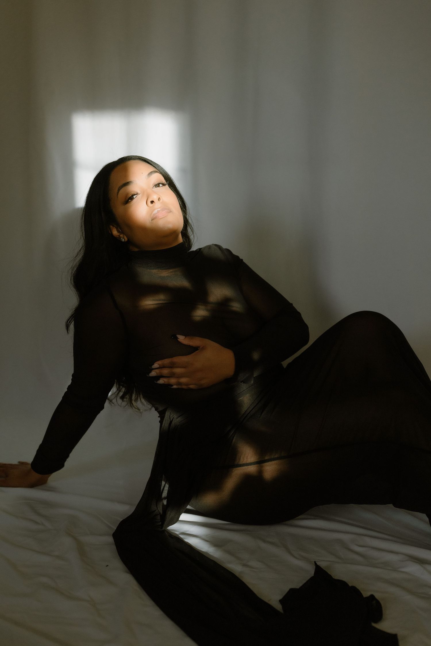 Woman in black turtleneck dress poses gracefully on white bed with dramatic window light casting shadows.