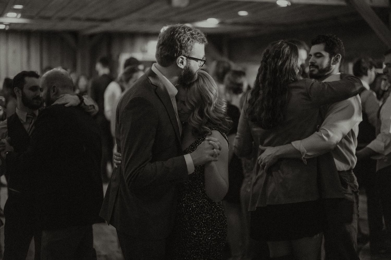 Couples slow dancing together at a wedding reception in a dimly lit venue, captured in black and white.