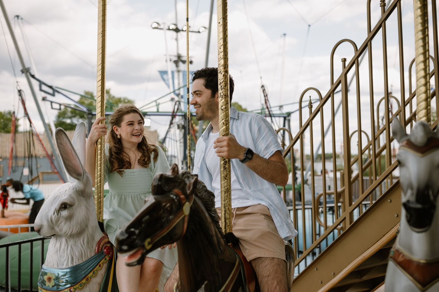Couple laughing and riding a carousel at an amusement park on colorful horse figures on a sunny day.