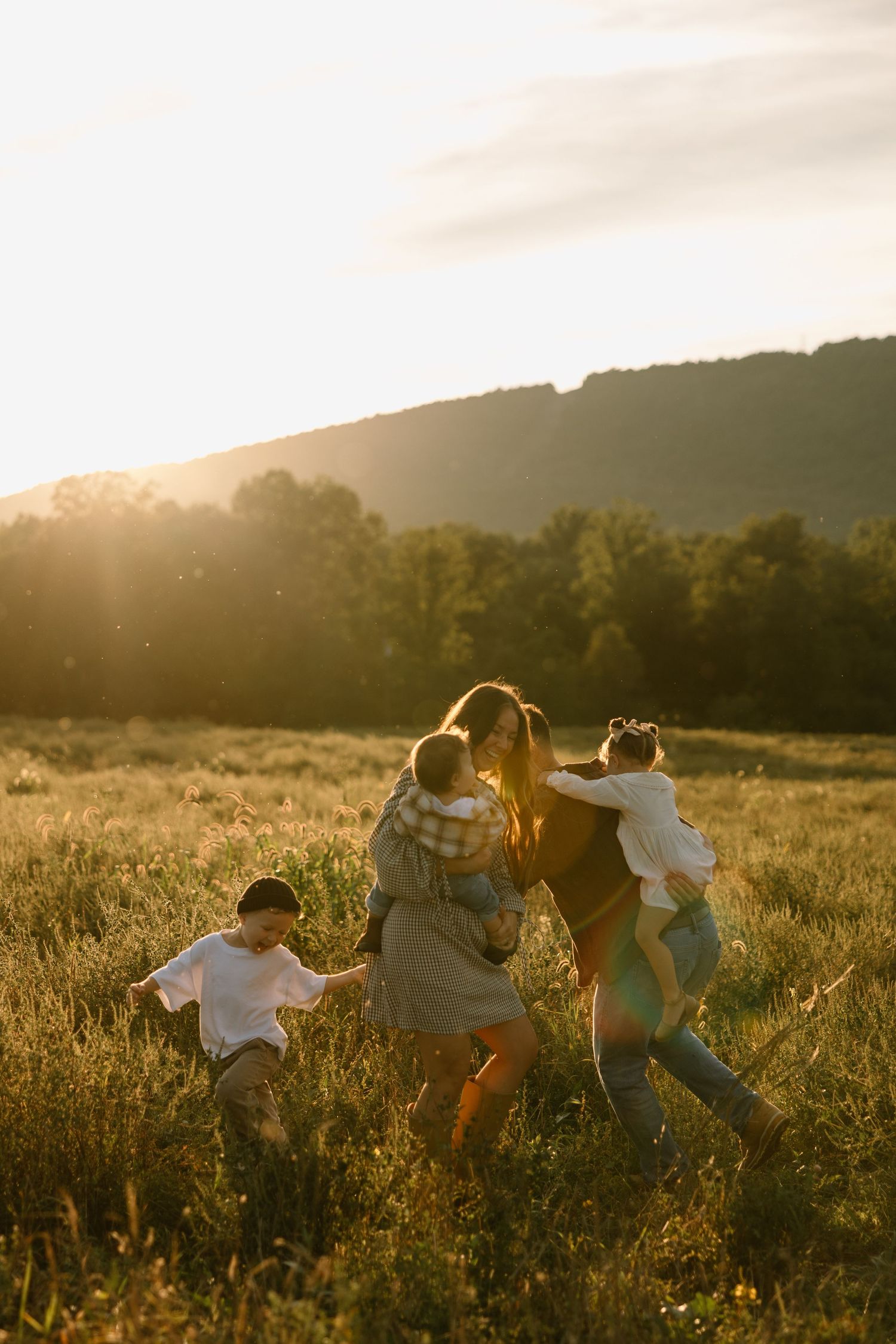 Family playing in a sunlit meadow at golden hour with mountains and trees in the background.