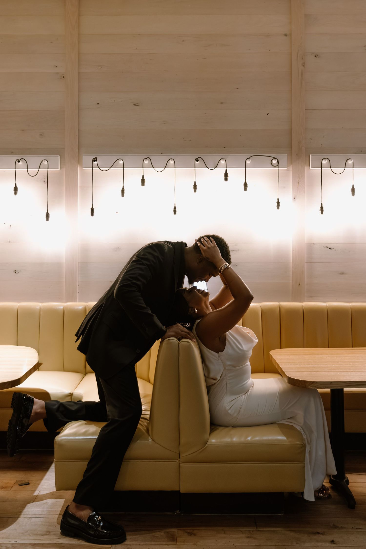 A couple shares a romantic moment in a cozy restaurant booth, man in black suit leaning to kiss woman in white dress.