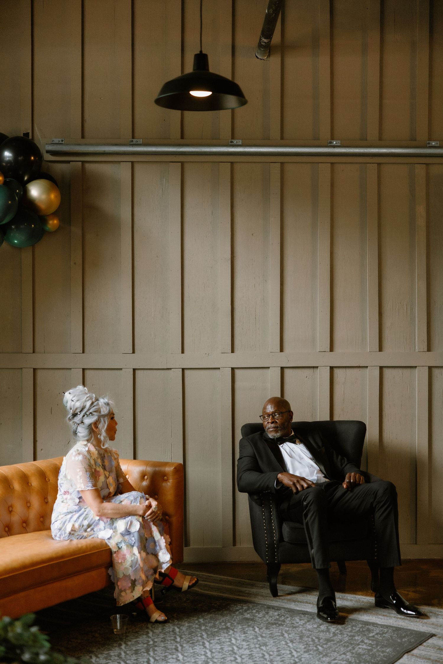 Two people in formal attire converse in a stylish lounge with wood-paneled walls, leather sofa, and balloon decor.