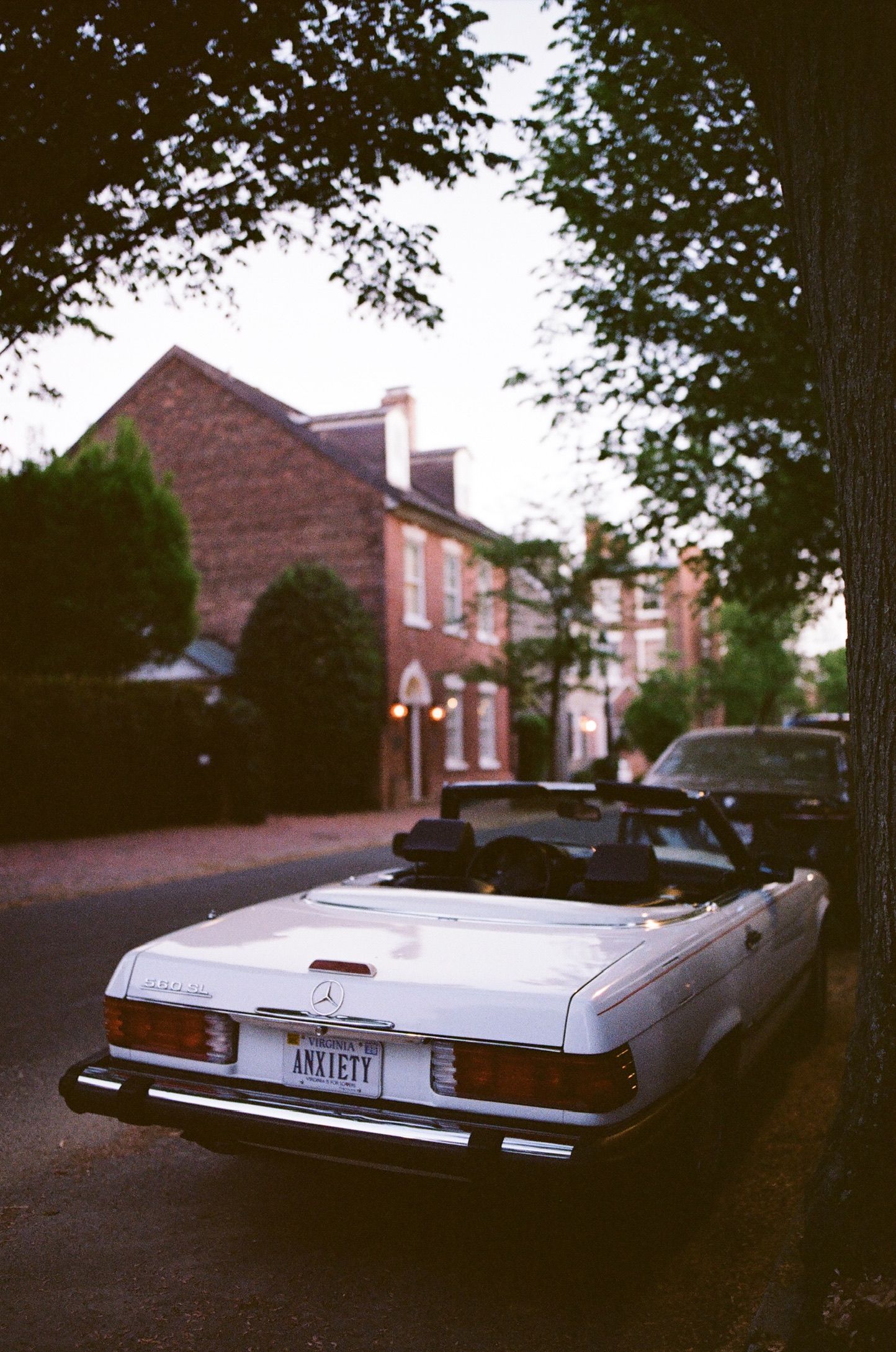 Vintage white Mercedes-Benz convertible with ANXIETY license plate parked on a tree-lined brick street near historic buildings.