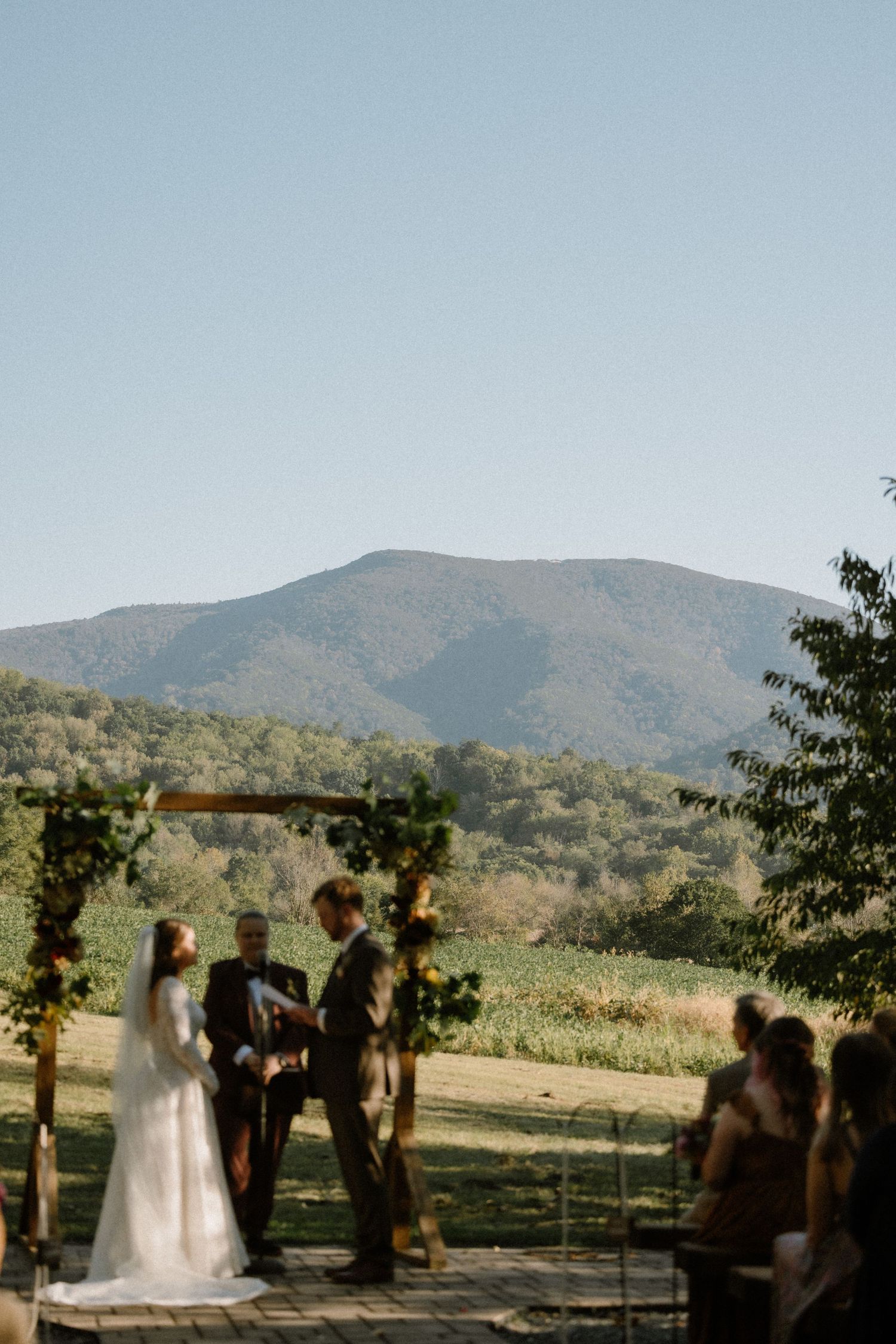 Outdoor wedding ceremony with bride and groom at floral arch, scenic mountain and green valley backdrop under clear blue sky.