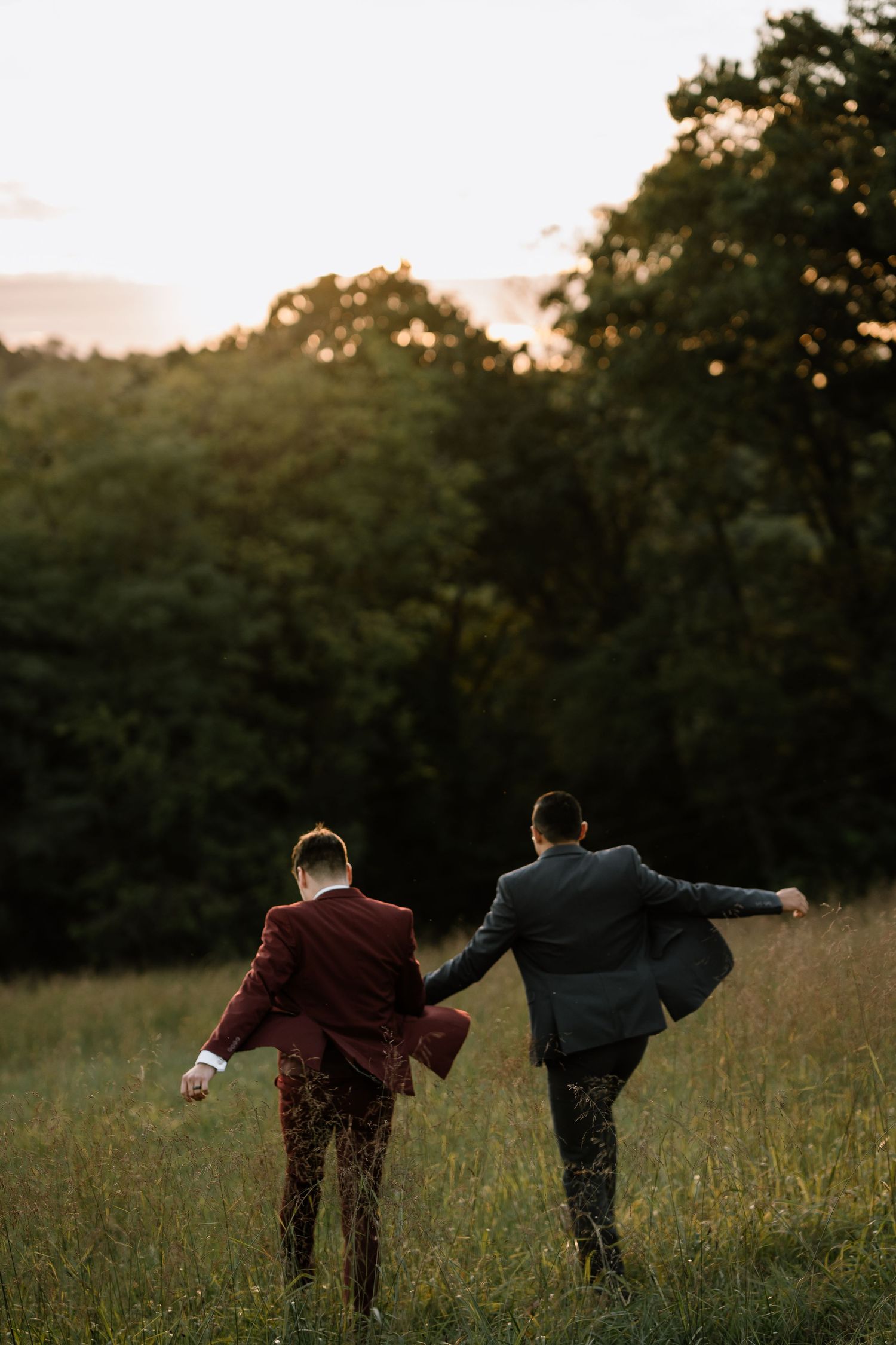 Two grooms in suits hold hands walking through a green meadow at sunset with trees in the background.