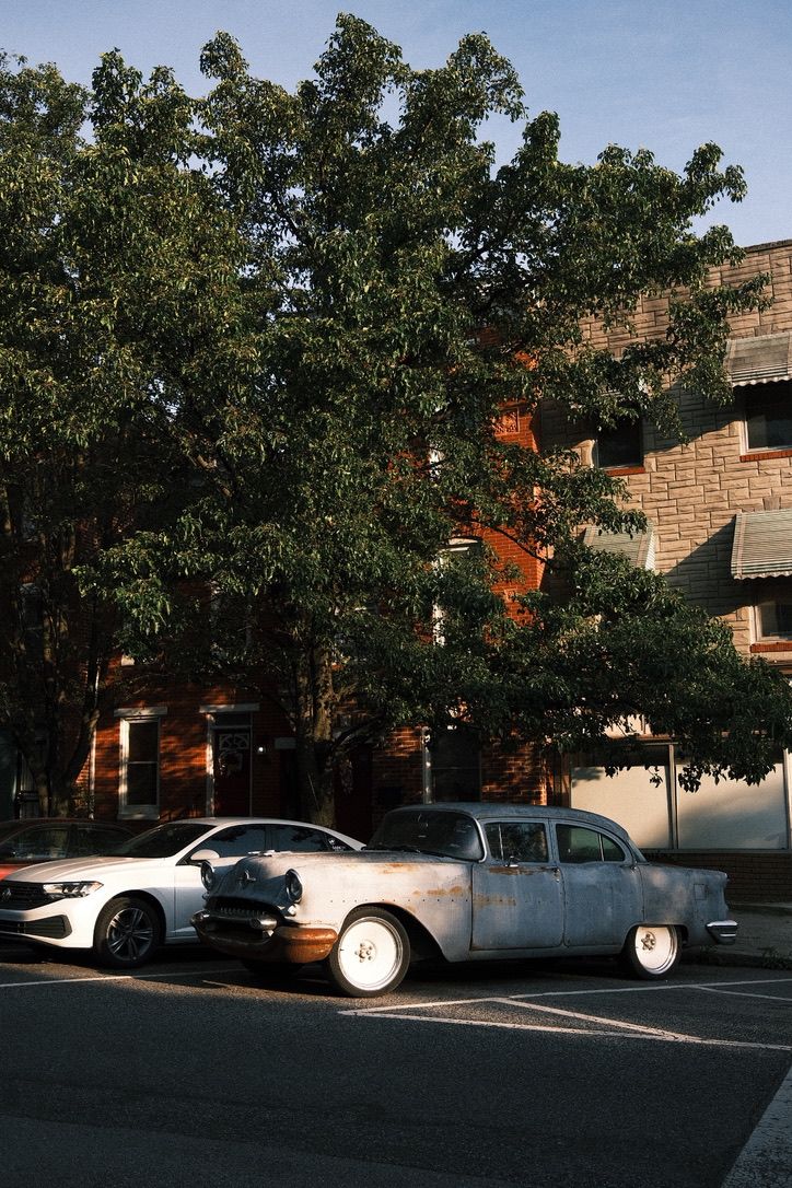 Vintage rusty classic car parked beside a modern white sedan near a brick building with large green trees in golden hour light.